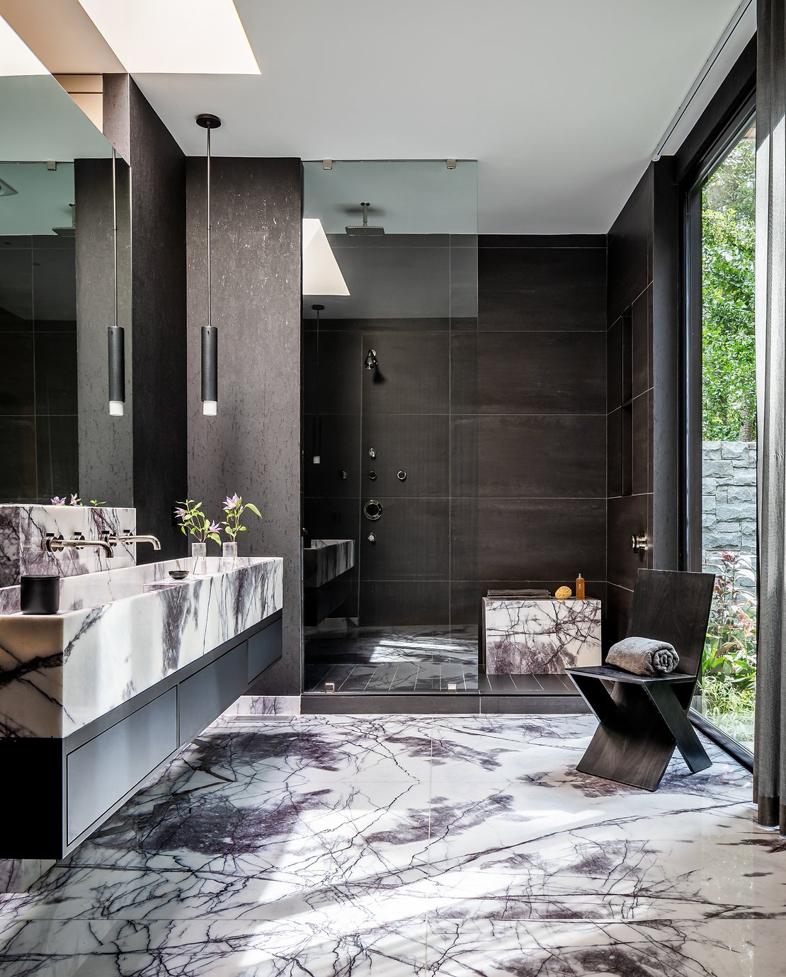 Custom primary bathroom with floating marble vanity, marble floor, dark tile shower, large mirror, pendant lights, and a wooden chair in Atlanta, GA.
