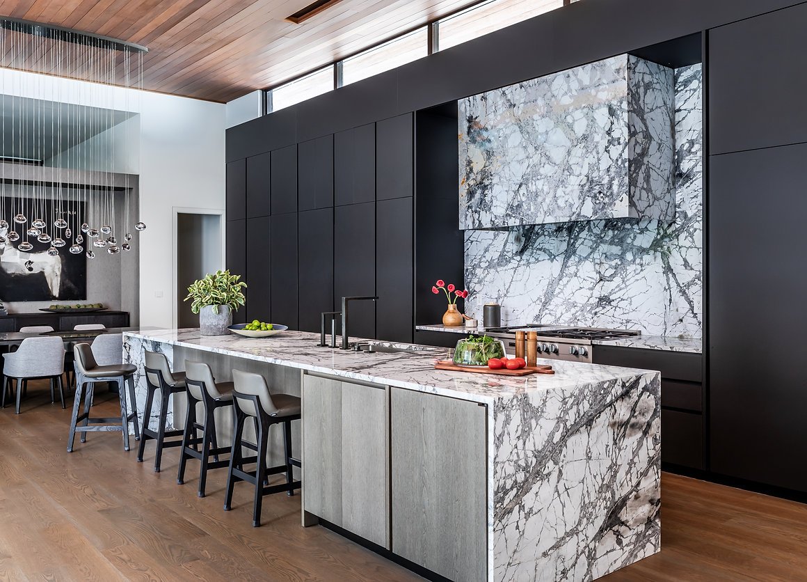 Modern kitchen with marble countertops, a large island, black cabinets, and wooden flooring. The kitchen features a sleek design with a dining area in the background and decorative lighting in Atlanta, GA.