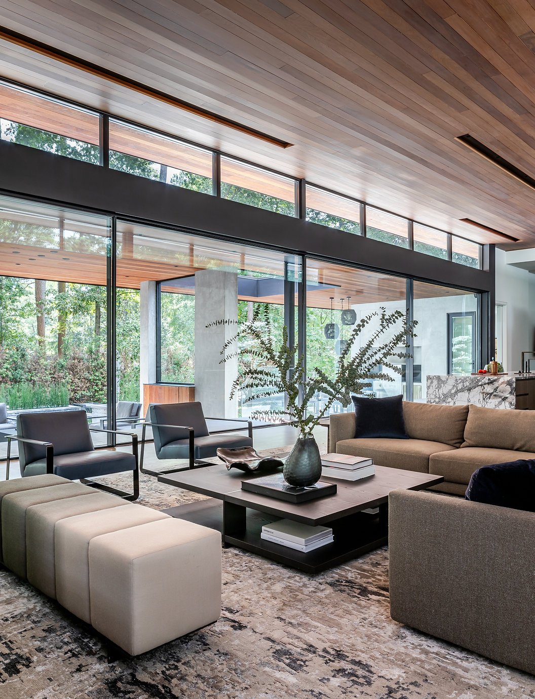 New construction of a modern living room with large windows, wooden ceiling, beige and brown Poliform sofa, a dark Poliform coffee table with a vase of greenery, and contemporary chairs, and custom ombre bench in Atlanta, GA on Old Ivy Road.