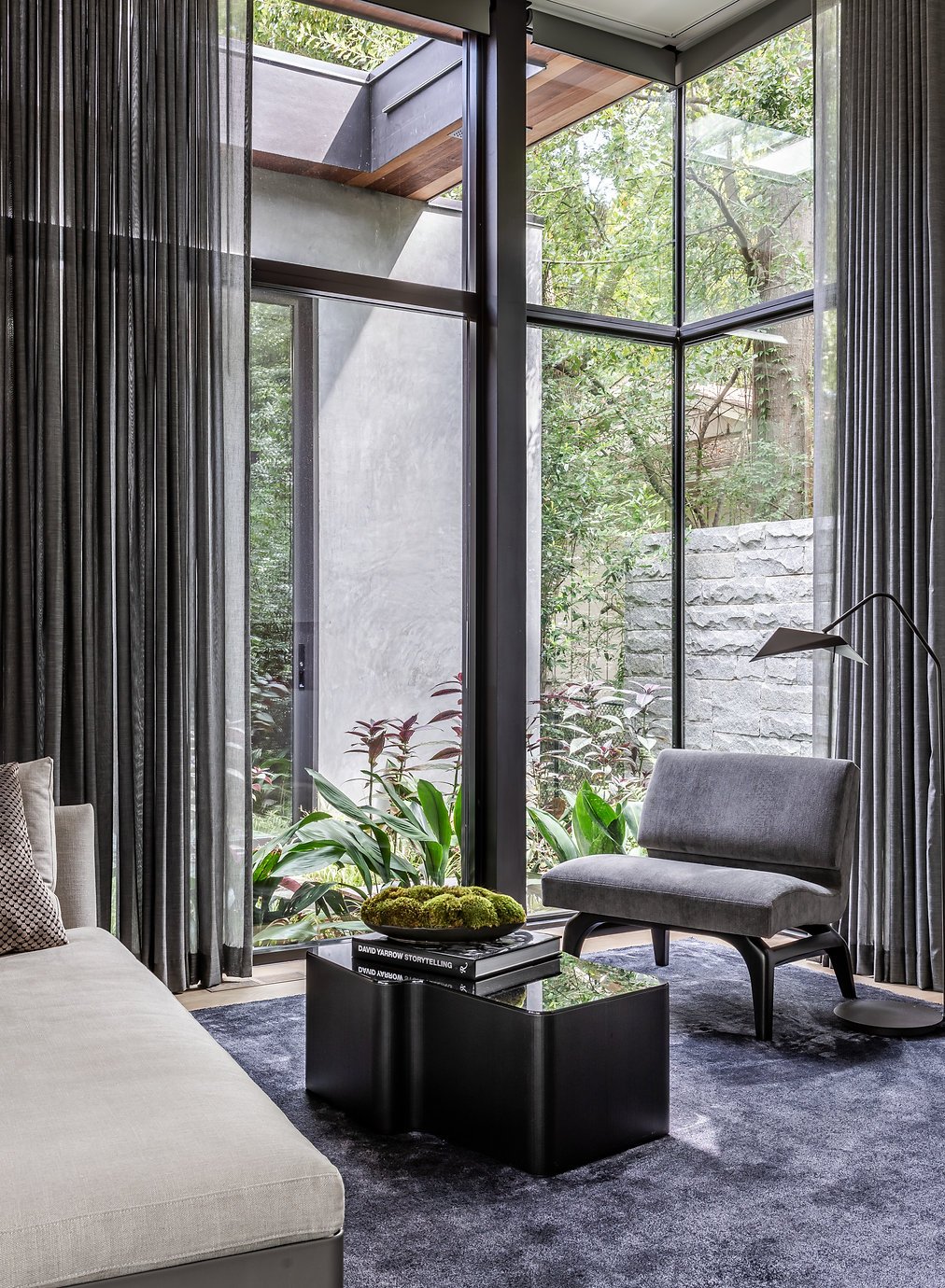 Modern primary sitting room with large windows, gray furniture, a Flexform chaise, a gray armchair, and a black coffee table overlooking an outdoor shower. The room features tall curtains, potted plants, and a view of greenery outside in Atlanta, GA.