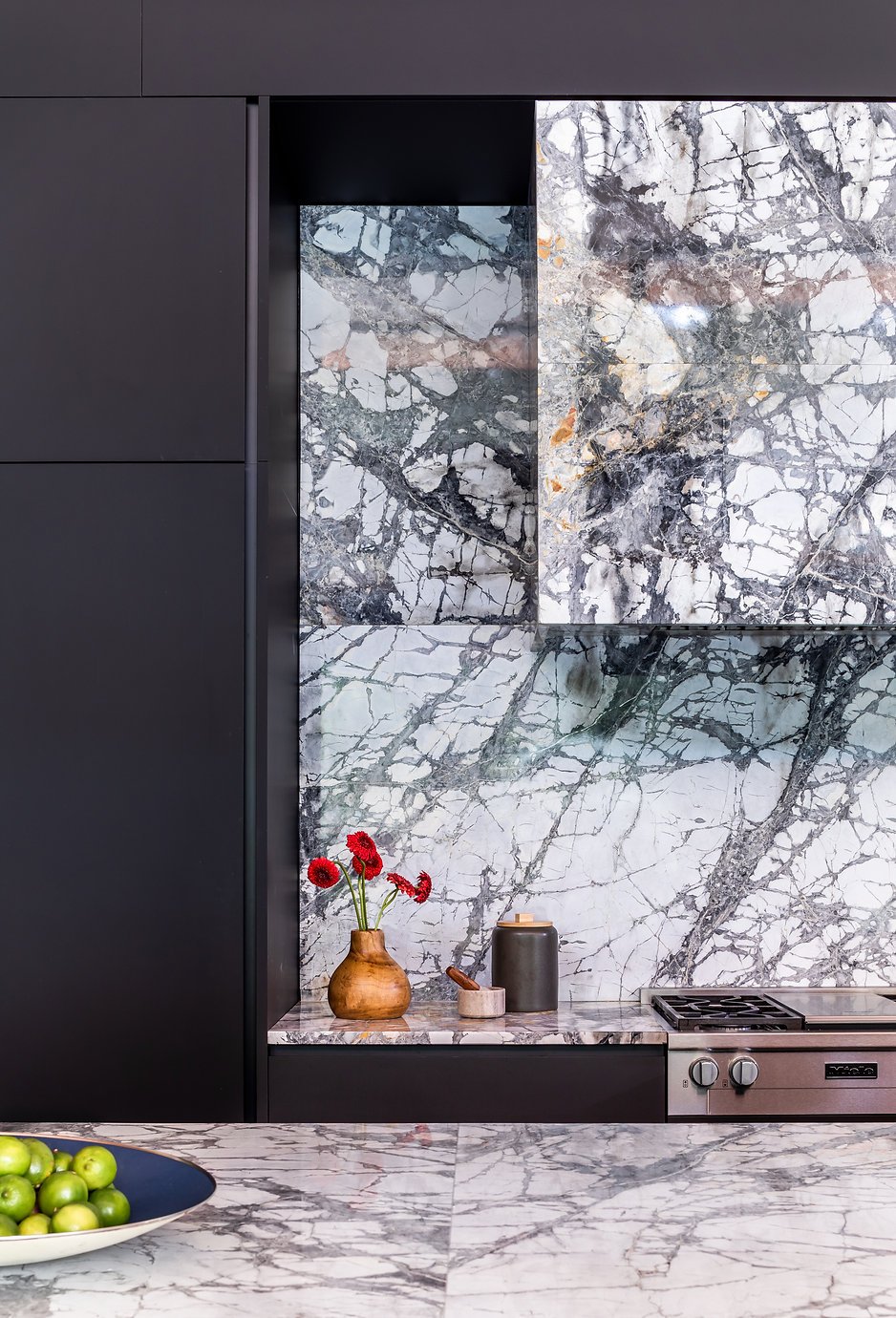 Modern kitchen with marble backsplash, black cabinets, a wooden vase with red flowers, a black canister, and a bowl of green apples on the marble countertop in Atlanta, GA.
