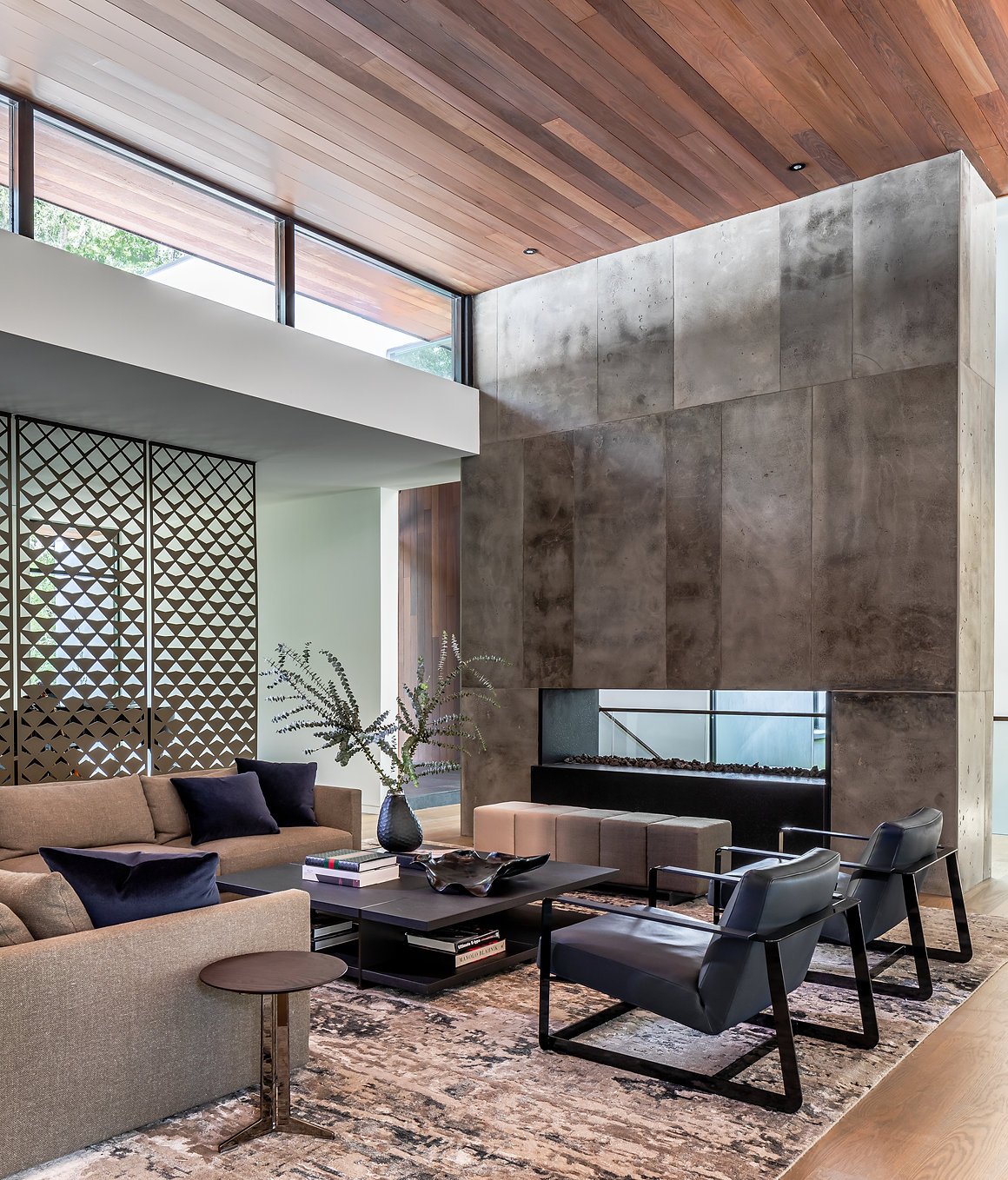 Modern living room with a beige Poliform sofa, dark armchairs, decorative vase with plants, custom ombre bench upholstered in Rosemary Halgarten fabric by Grant Trick, large concrete fireplace, and wooden ceiling in Atlanta, GA.
