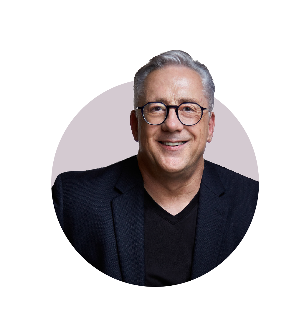 A smiling middle-aged man with gray hair, glasses, wearing a black blazer and black shirt against a purple and white abstract background.
