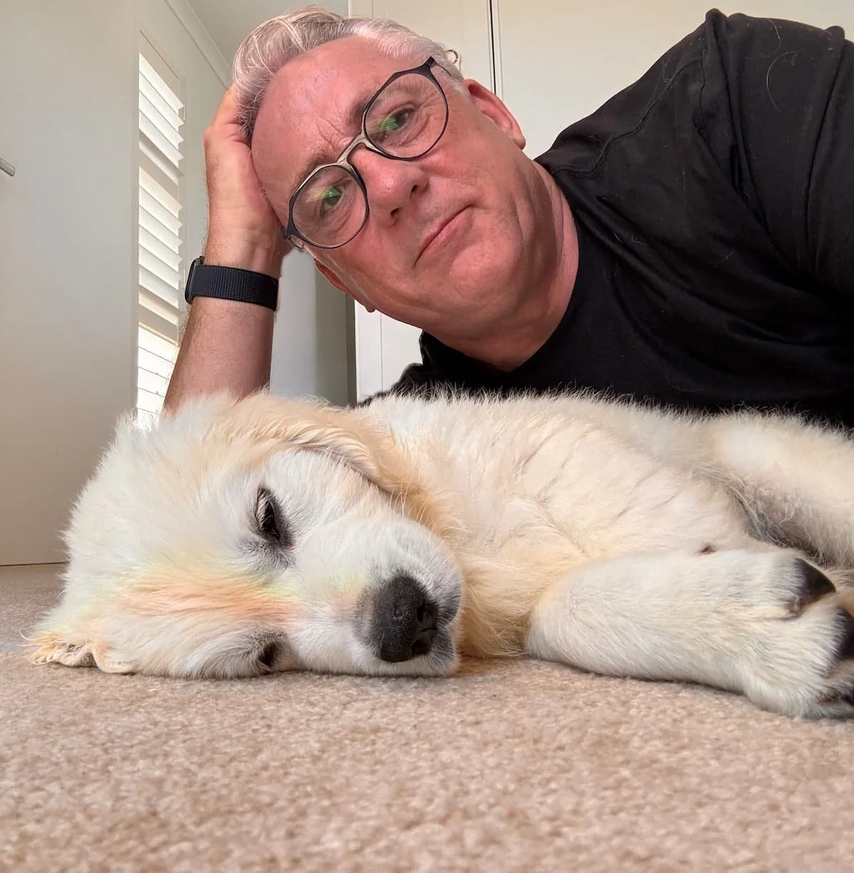 A man with glasses and a black shirt laying next to a sleeping golden retriever puppy on a carpeted floor in a room with white walls and a window with blinds.