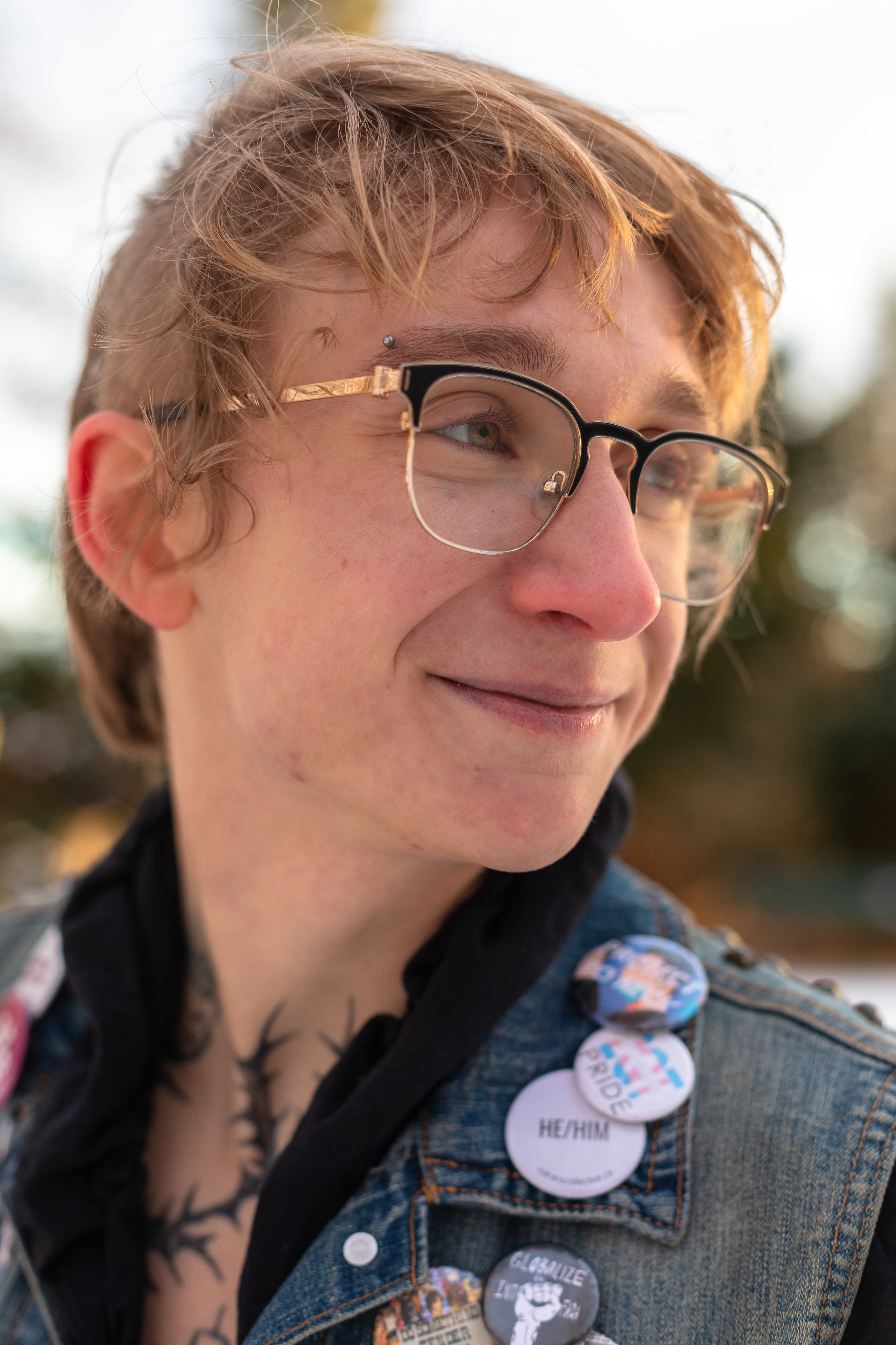 Close-up of a person with light brown tousled hair, wearing glasses and a denim vest adorned with buttons, smiling outdoors in natural light.