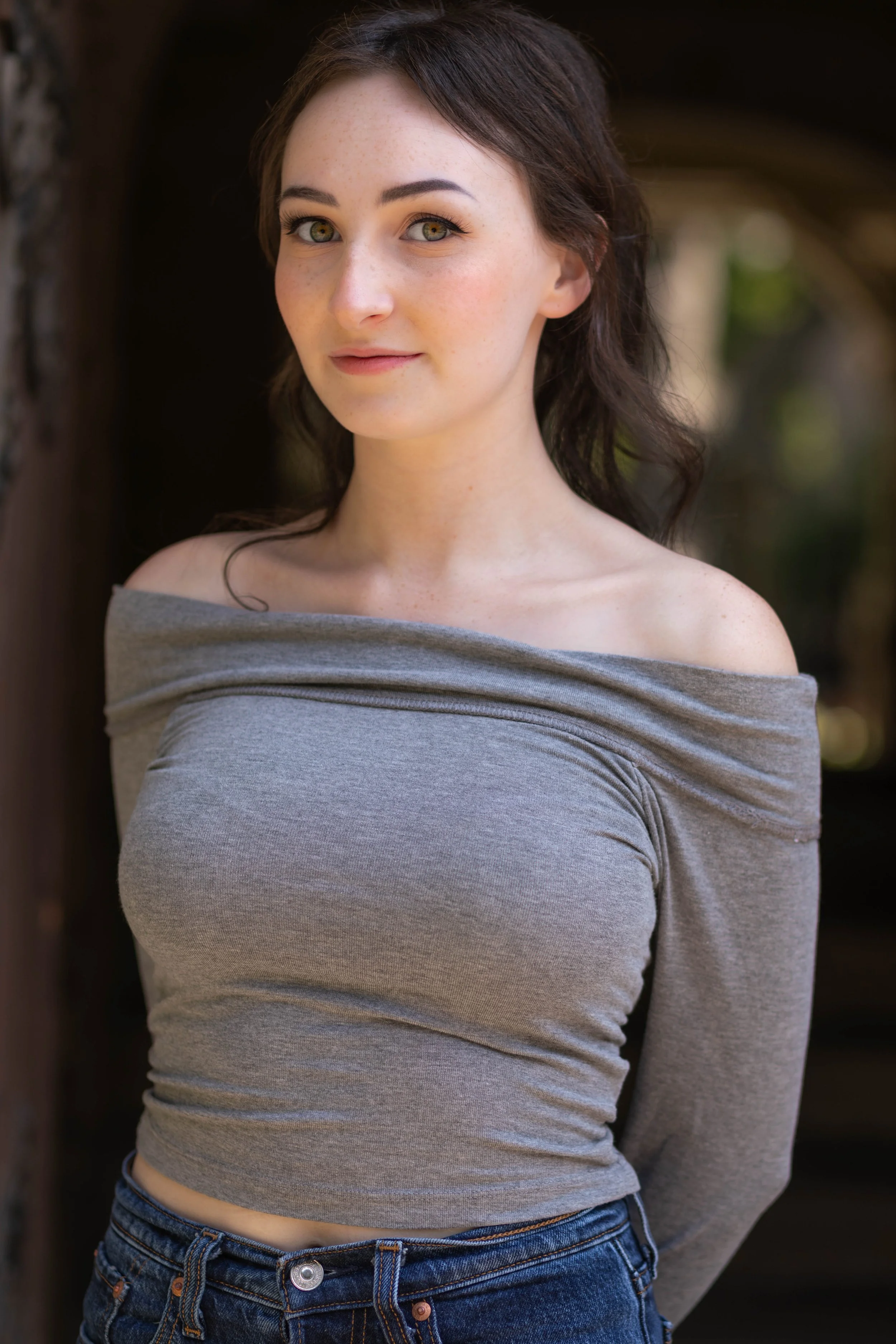A young woman with fair skin, freckles, dark brown hair, and yellow-green eyes, standing outdoors, wearing an off-shoulder gray top and blue jeans.