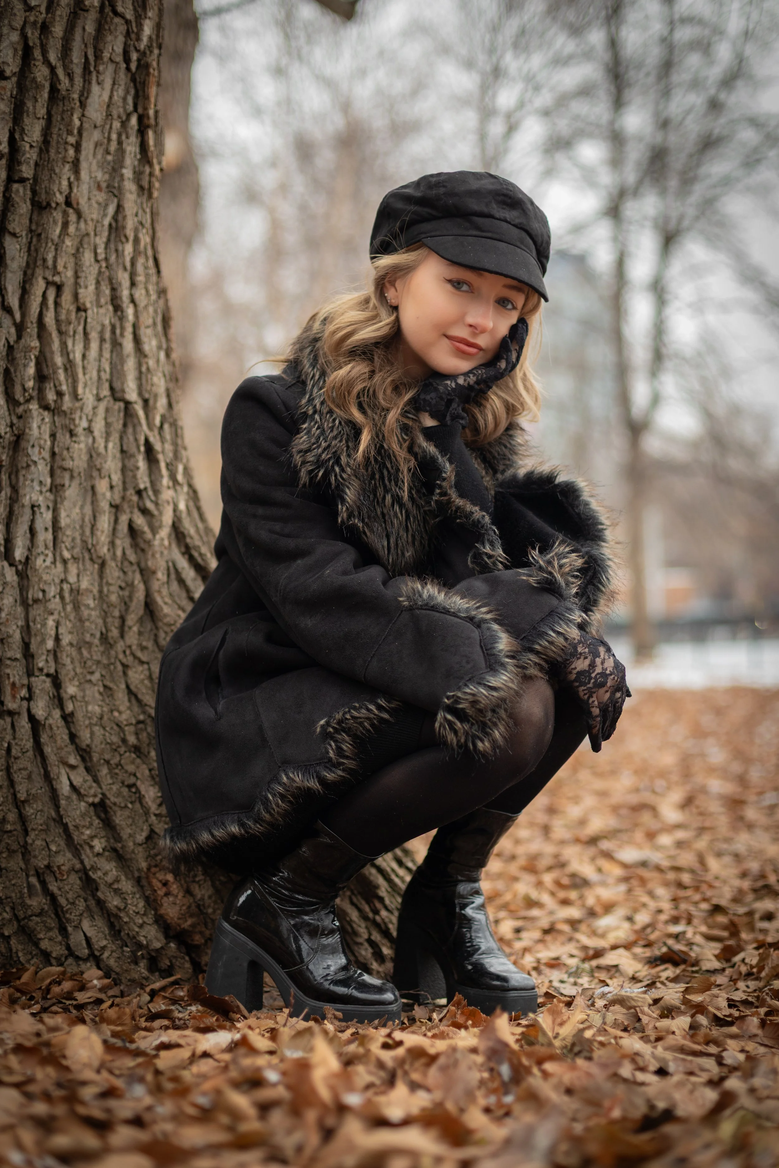 A woman crouching beside a tree trunk in a park during autumn, wearing a black coat with fur trim, a black hat, lace gloves, and shiny black high heels. She has wavy blonde hair and a subtle smile.