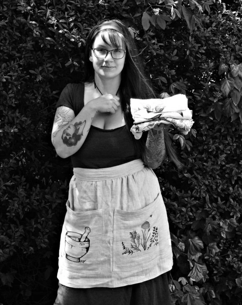 A young woman with long hair and glasses standing outdoors in front of foliage, holding a stack of folded cloths or towels in her right hand and resting her left hand on her chest, wearing a black top and a light apron with embroidered pockets.