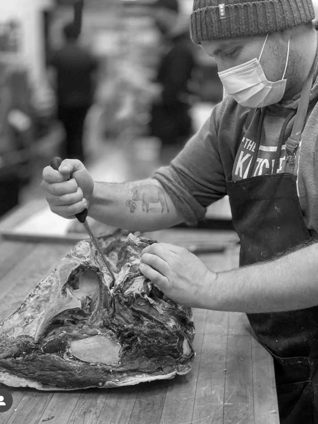 A man wearing a beanie, face mask, and apron but no gloves, is carving a large piece of meat on a wooden surface in a busy environment, possibly a butcher shop or kitchen.