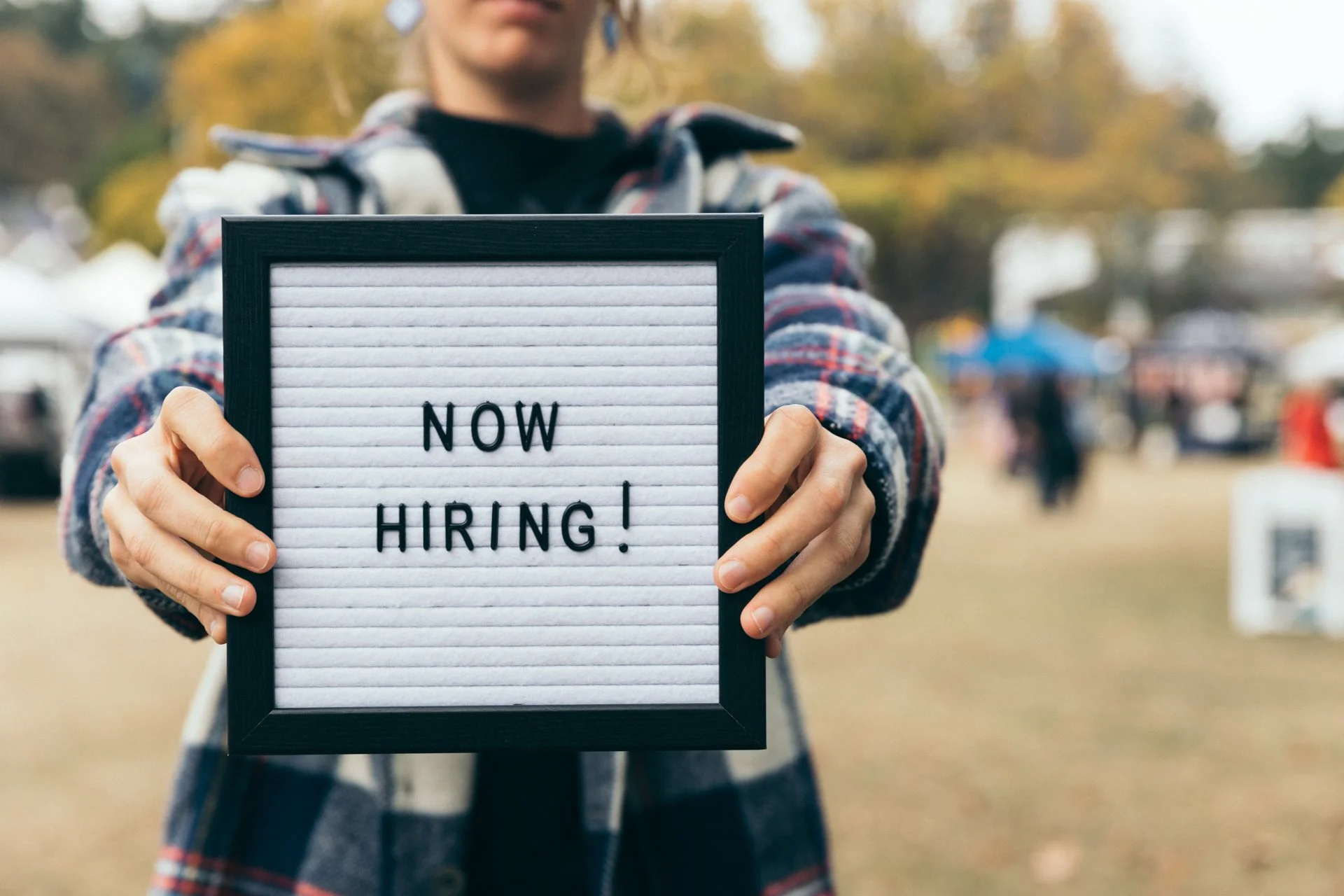 Person holding a letter board with the message 'NOW HIRING!' at the Esquimalt Farmers Market, with a blurred background of trees and people.