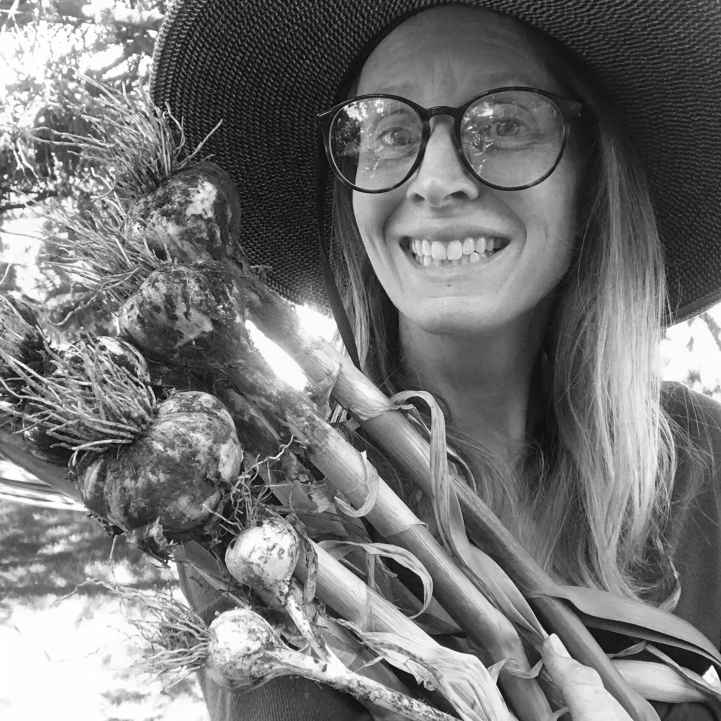 A woman smiling, wearing glasses and a wide-brimmed hat, holding freshly harvested carrots.