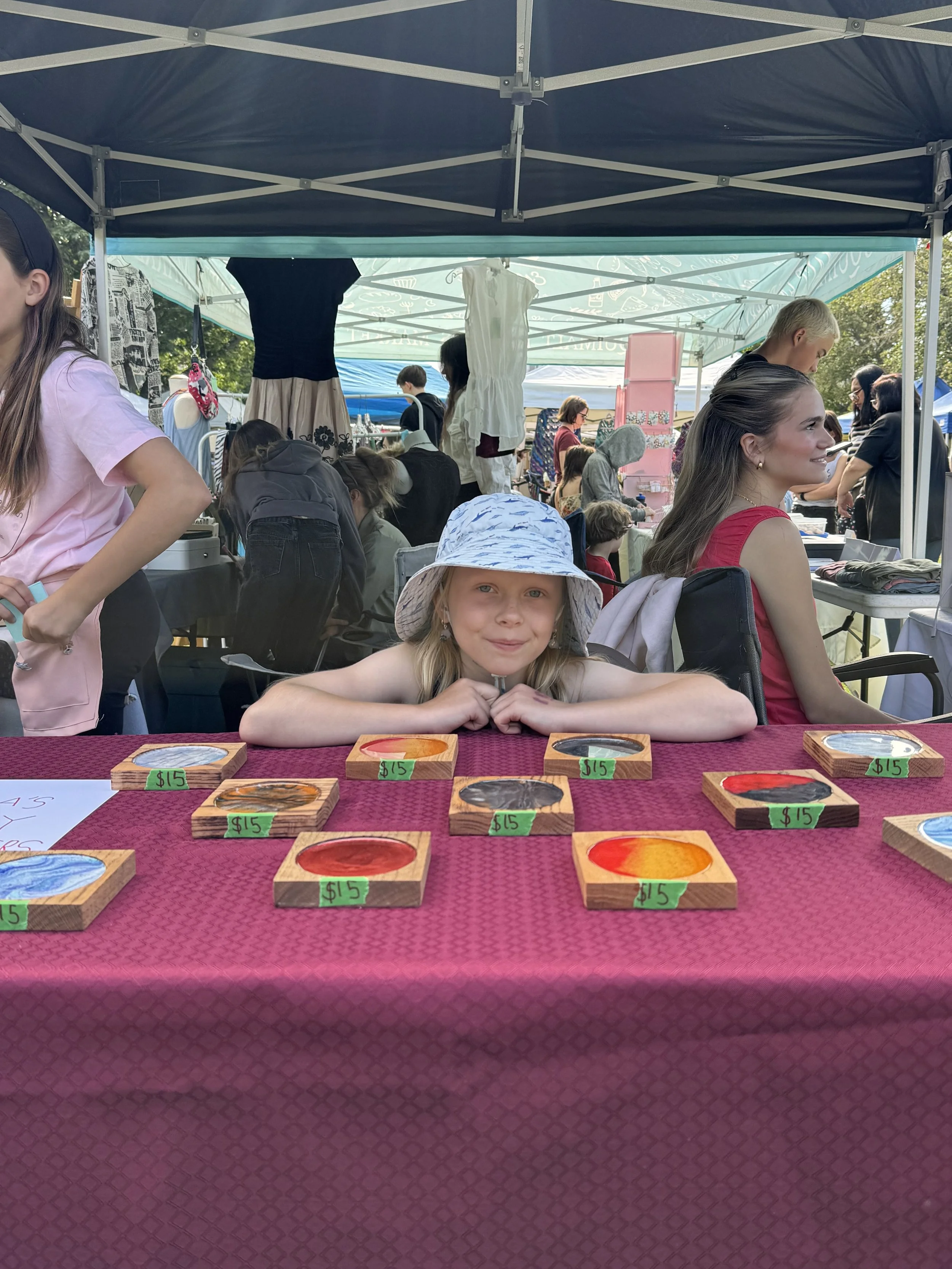 A girl with blonde hair and a sunhat sitting at a table with painted wooden coasters, each priced at $15, under a tent at an outdoor market.