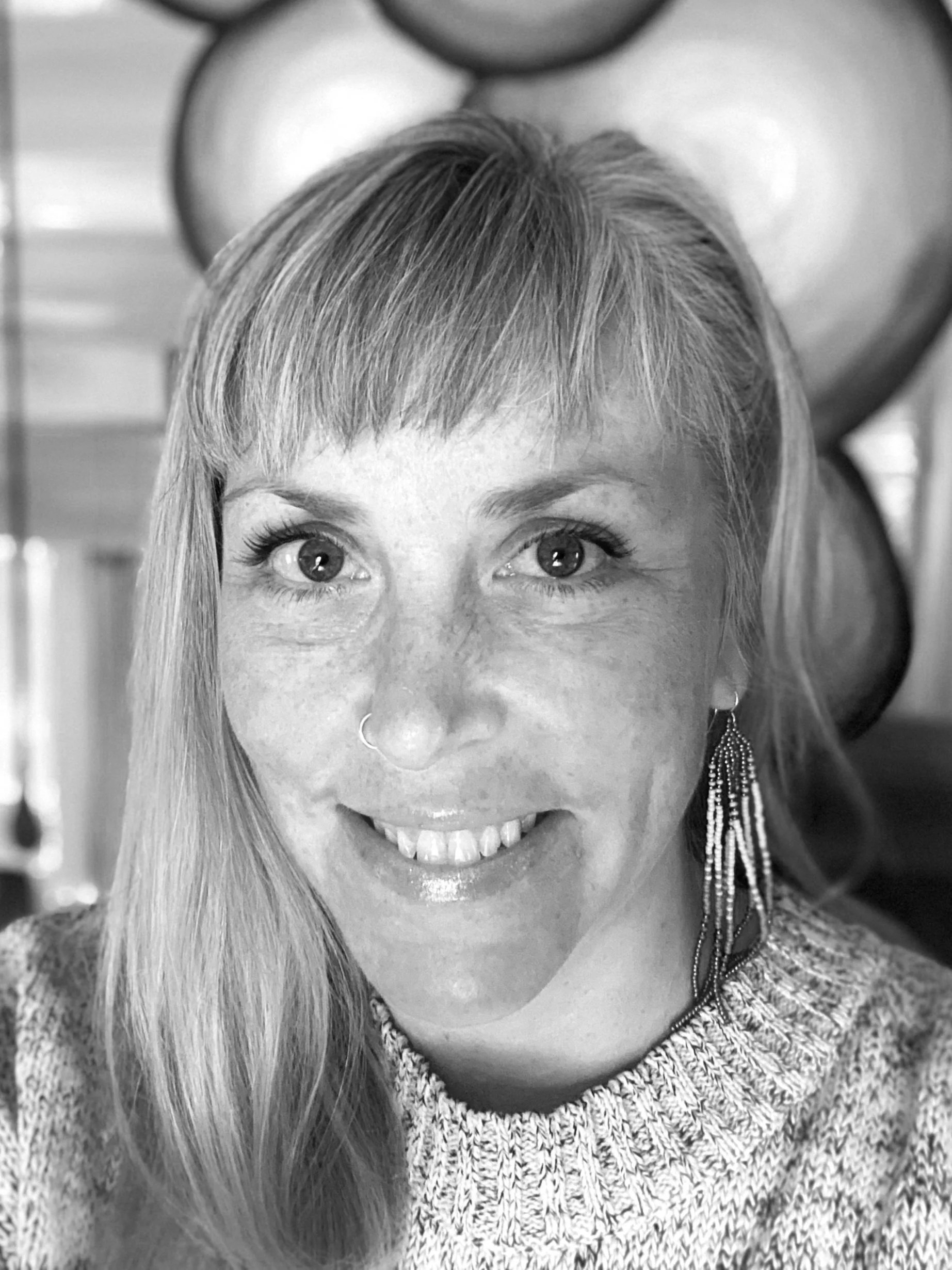 Black and white close-up photo of a smiling woman with short hair, wearing earrings and a knitted sweater, with a background that includes a large heart-shaped decor.