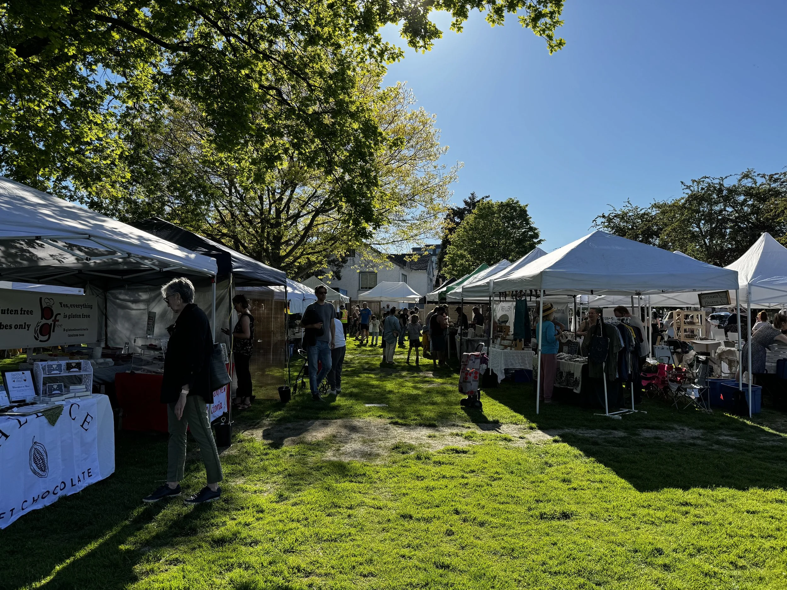 The outdoor Esquimalt Farmers Market featuring white tents and people walking on grass, surrounded by trees, under a clear blue sky.