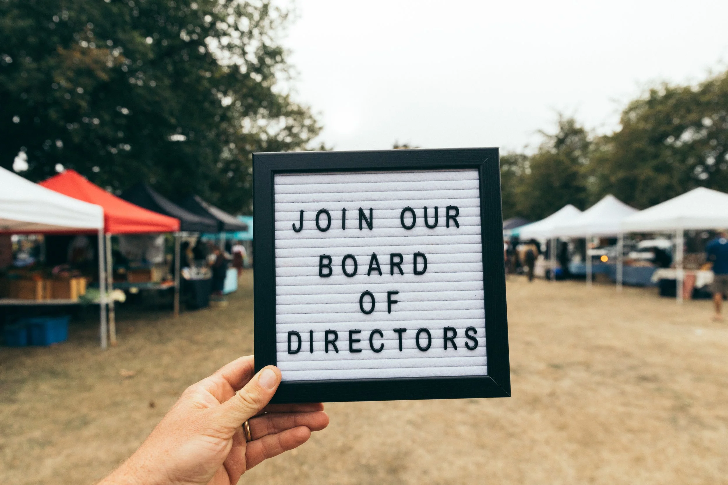 A person holding a letter board with the words 'JOIN OUR BOARD OF DIRECTORS' displayed on it, outdoors at the Esquimalt Farmers Market with tents and trees in the background.