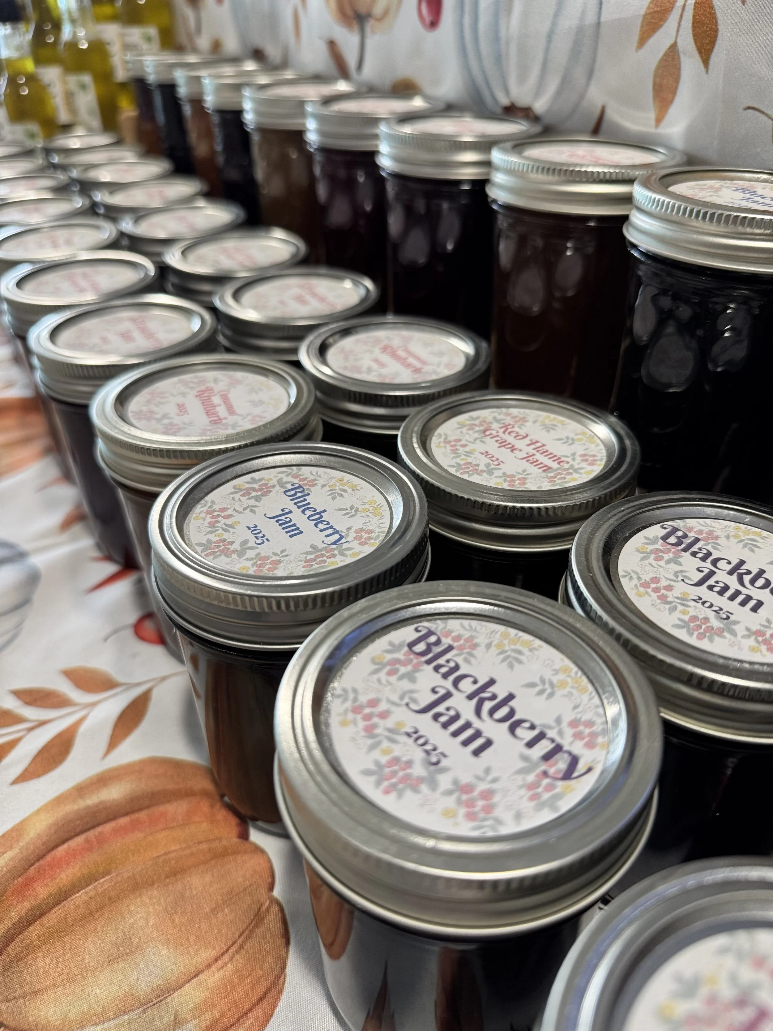 Several jars of homemade blackberry jam with metal lids, arranged on a table with a fall-themed tablecloth featuring pumpkins.