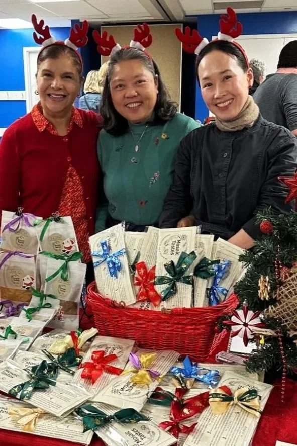 Three women wearing Christmas antler headbands, smiling, standing behind a table filled with wrapped gifts and informational leaflets at the Esquimalt Holiday Market.
