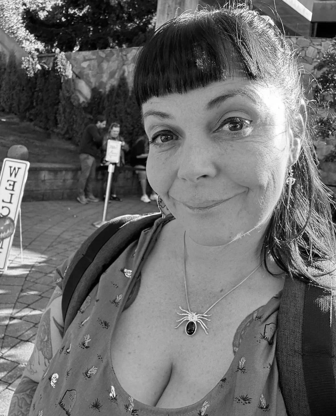 A woman with short dark hair and bangs takes a selfie outdoors, smiling gently. She wears a patterned top, a spider-shaped necklace with a black stone, and earrings. In the background, two people are standing near a small sign and a stone wall with trees behind.