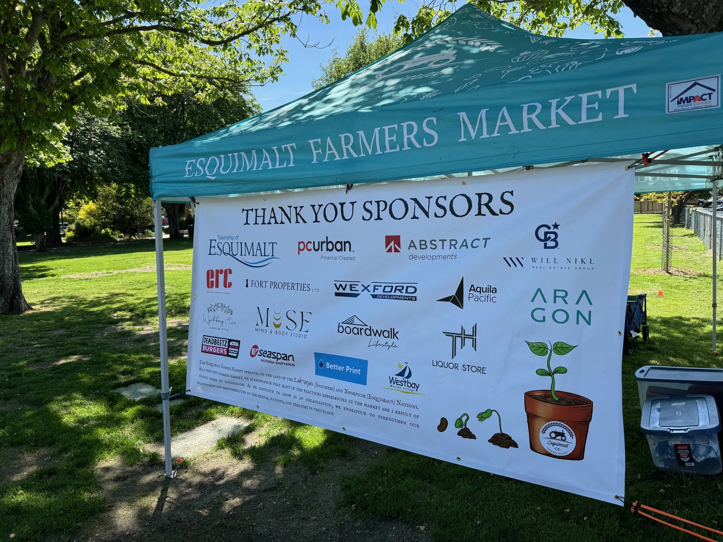 A large canopy with the words 'Esquimalt Farmers Market' printed on it, set up outdoors among trees and grass. Below the canopy, there is a banner thanking sponsors with logos and names of various organizations, and a small cartoon illustration of a plant in a pot.