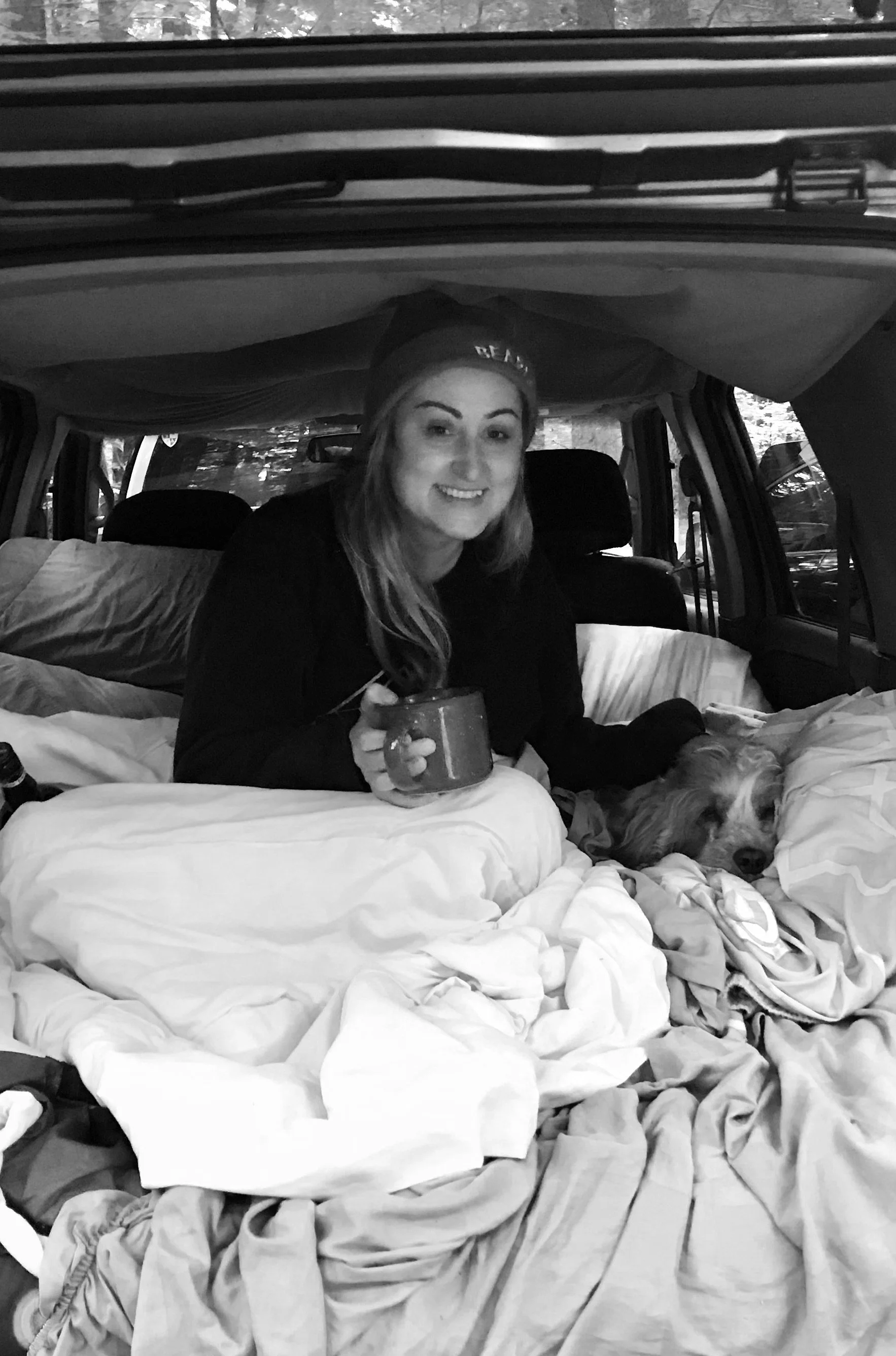 A woman with long hair, wearing a cap and dark jacket, smiling and holding a mug, sitting in the back of a vehicle with blankets and a sleeping dog beside her.