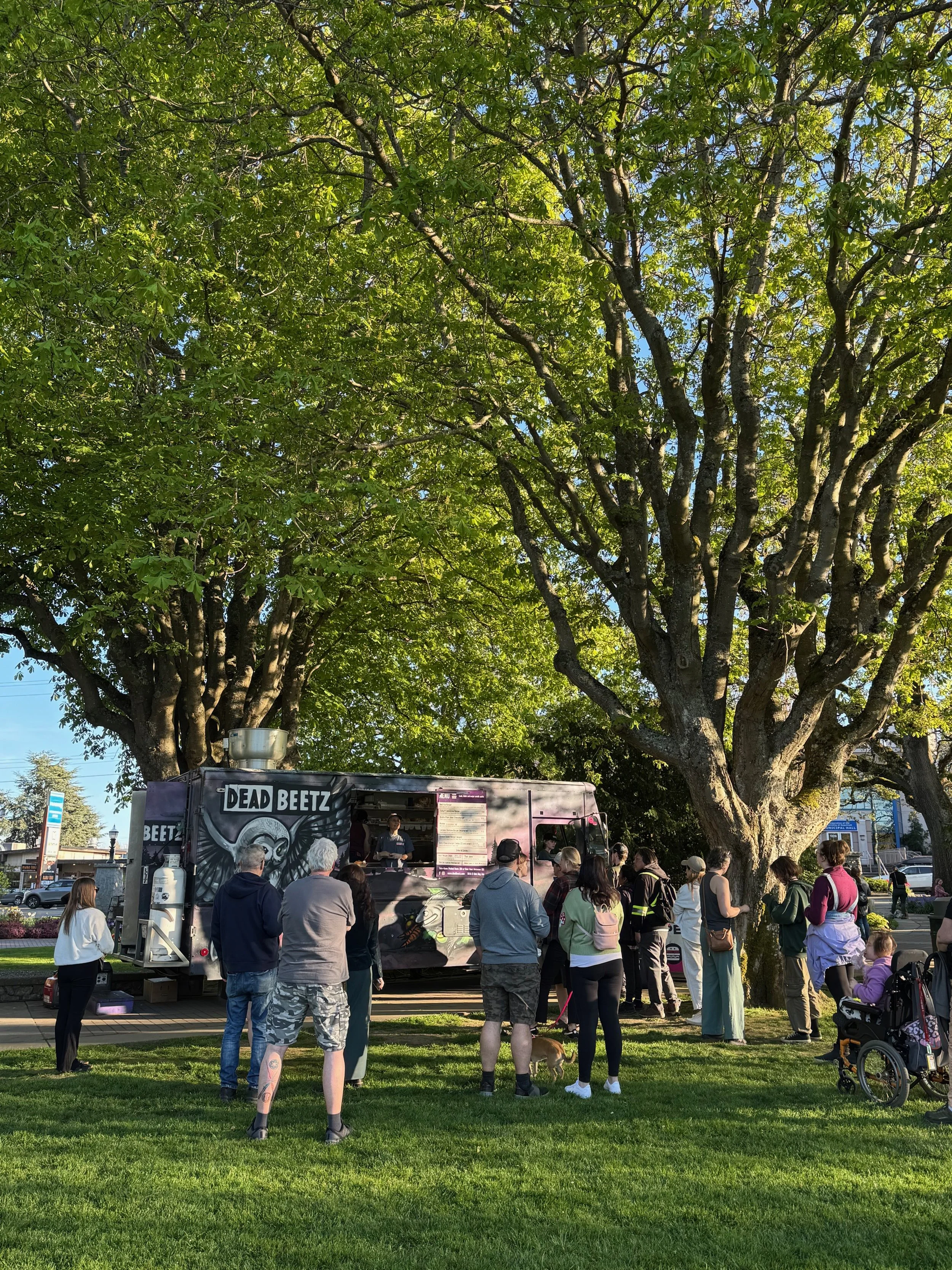A crowd of people at Esquimalt Farmers Market waiting in line at a food truck, called Dead Beetz, serving food under large trees in a park. The scene is sunny and outdoors.