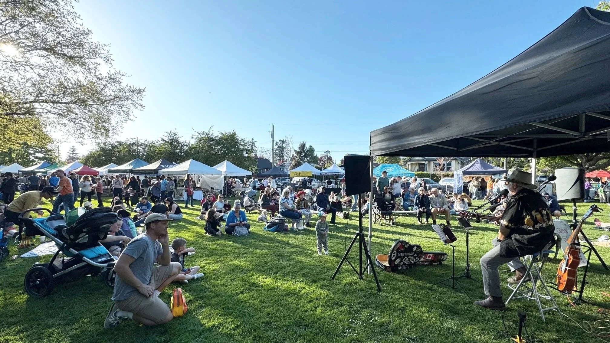 Outdoor community event, the Esquimalt Farmers Market with tents, people sitting on grass, a musician playing guitar, and a bright sunny day.