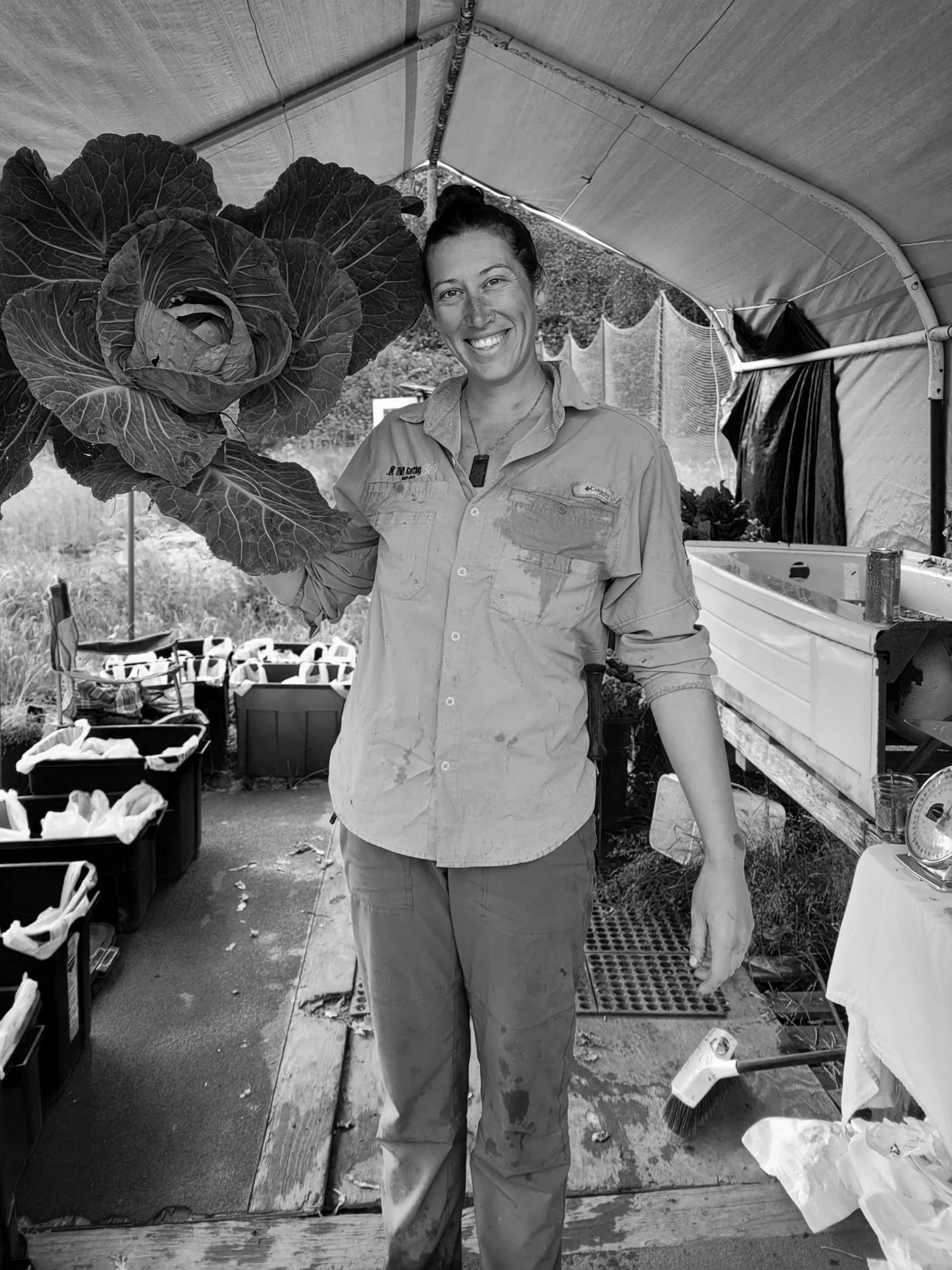 A woman holding a large head of lettuce, smiling, standing in a greenhouse or outdoor farm setup surrounded by gardening supplies and produce.