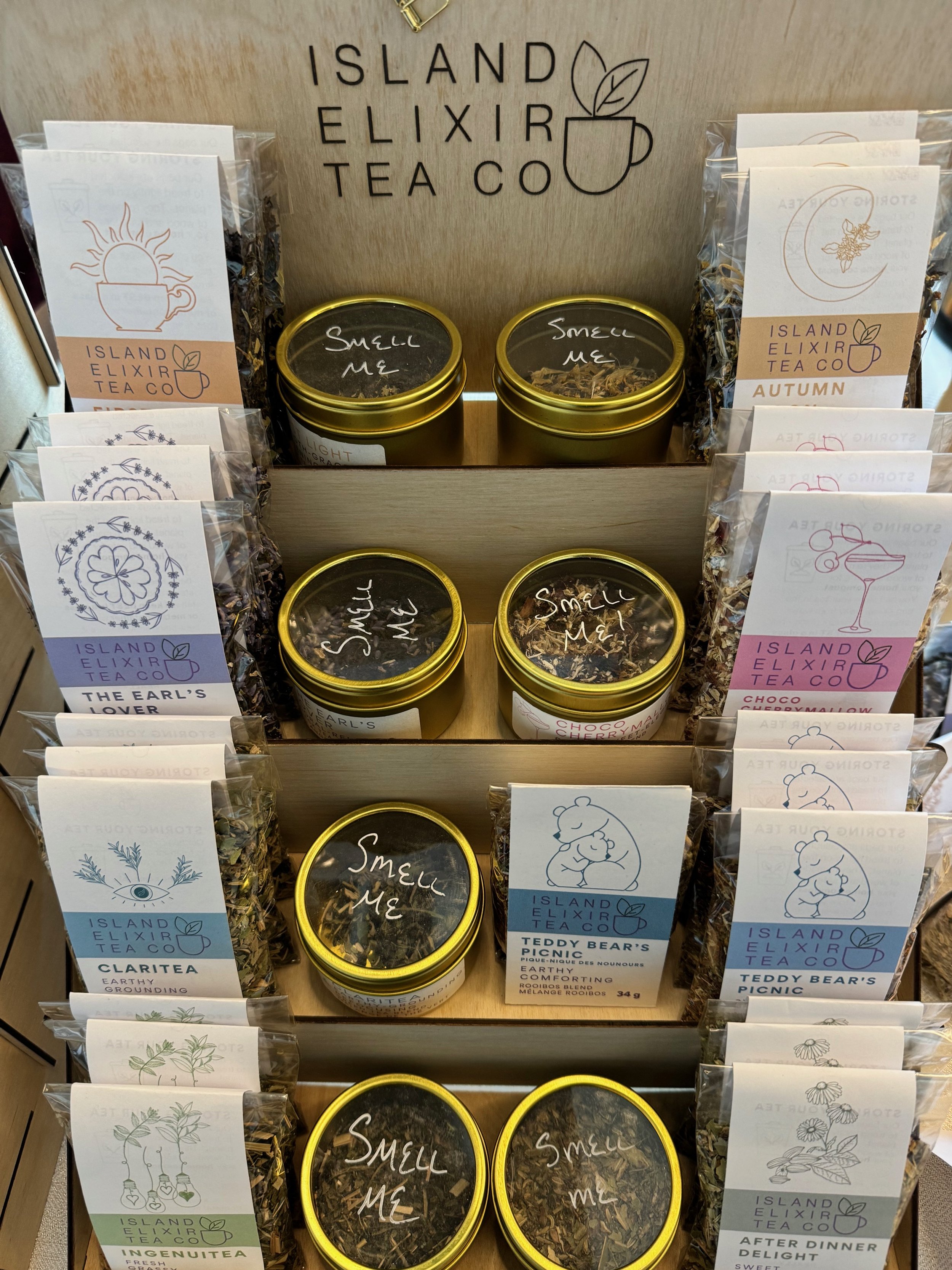 Display of various tins and packets of tea from Island Elixir Tea Co., with labels like 'Smell Me,' 'Claritea,' 'Teddy Bear's Picnic,' and 'After Dinner Delight,' arranged on shelves.