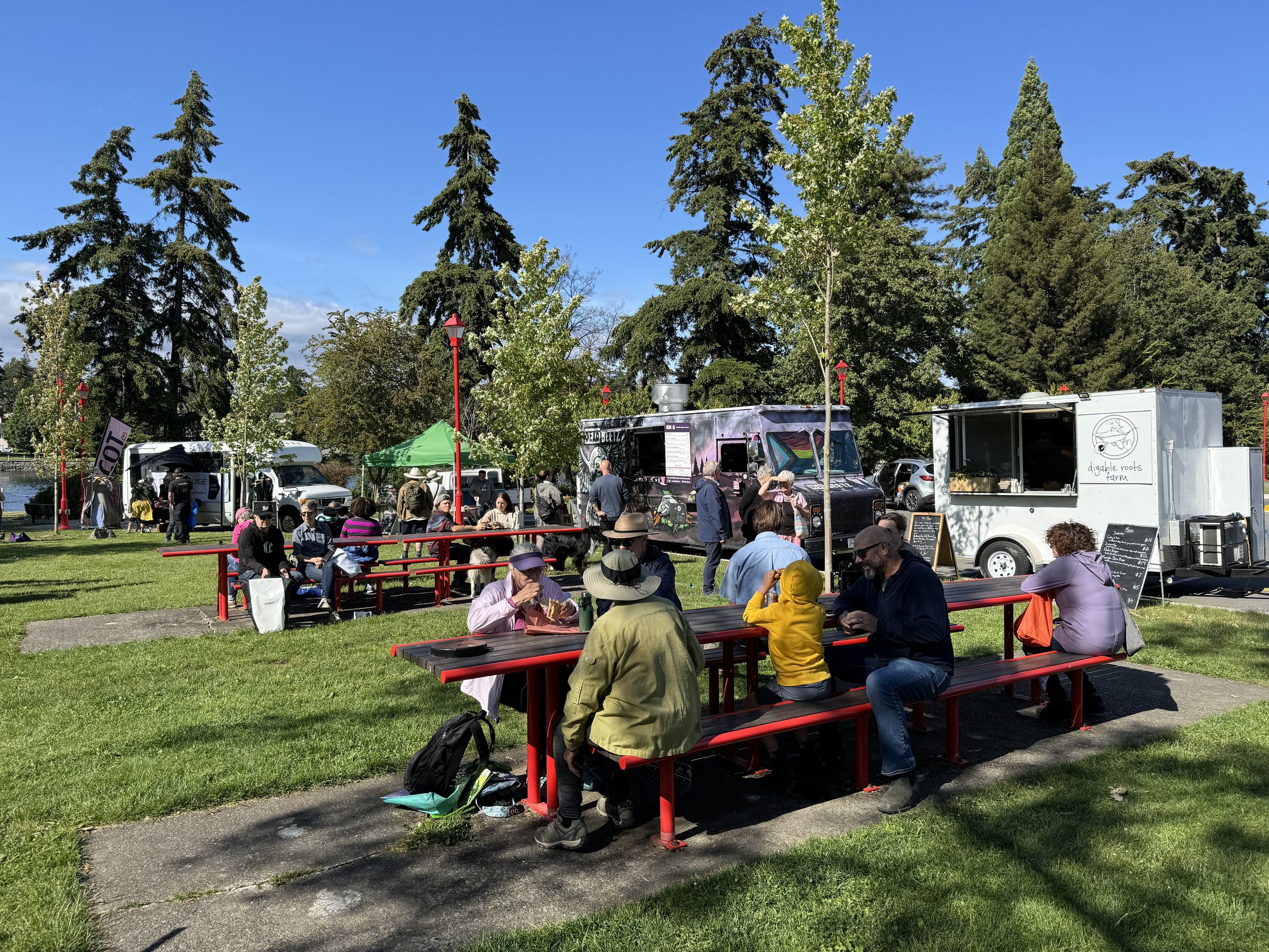 People gathered outdoors at a sunny farmers market, sitting on red benches and conversing, with food trucks and trees in the background.