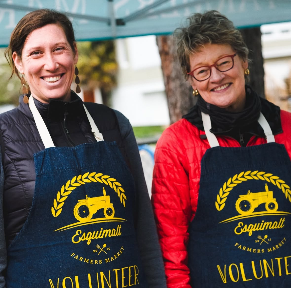 Two women smiling at a farmers market, wearing aprons with the Esquimalt Farmers Market logo that reads 'Volunteer'.