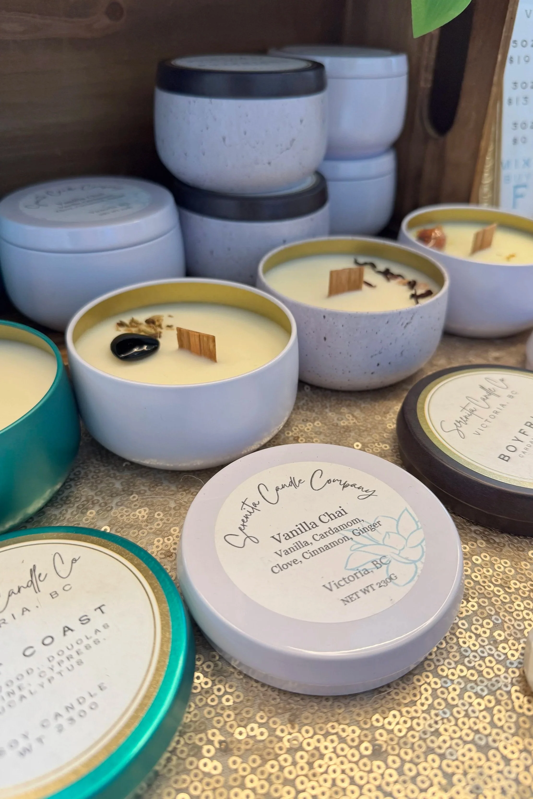 Several small candles in decorative containers with labels indicating flavors like vanilla chai, cinnamon, ginger, and clove, arranged on a decorative surface with a wooden backdrop.