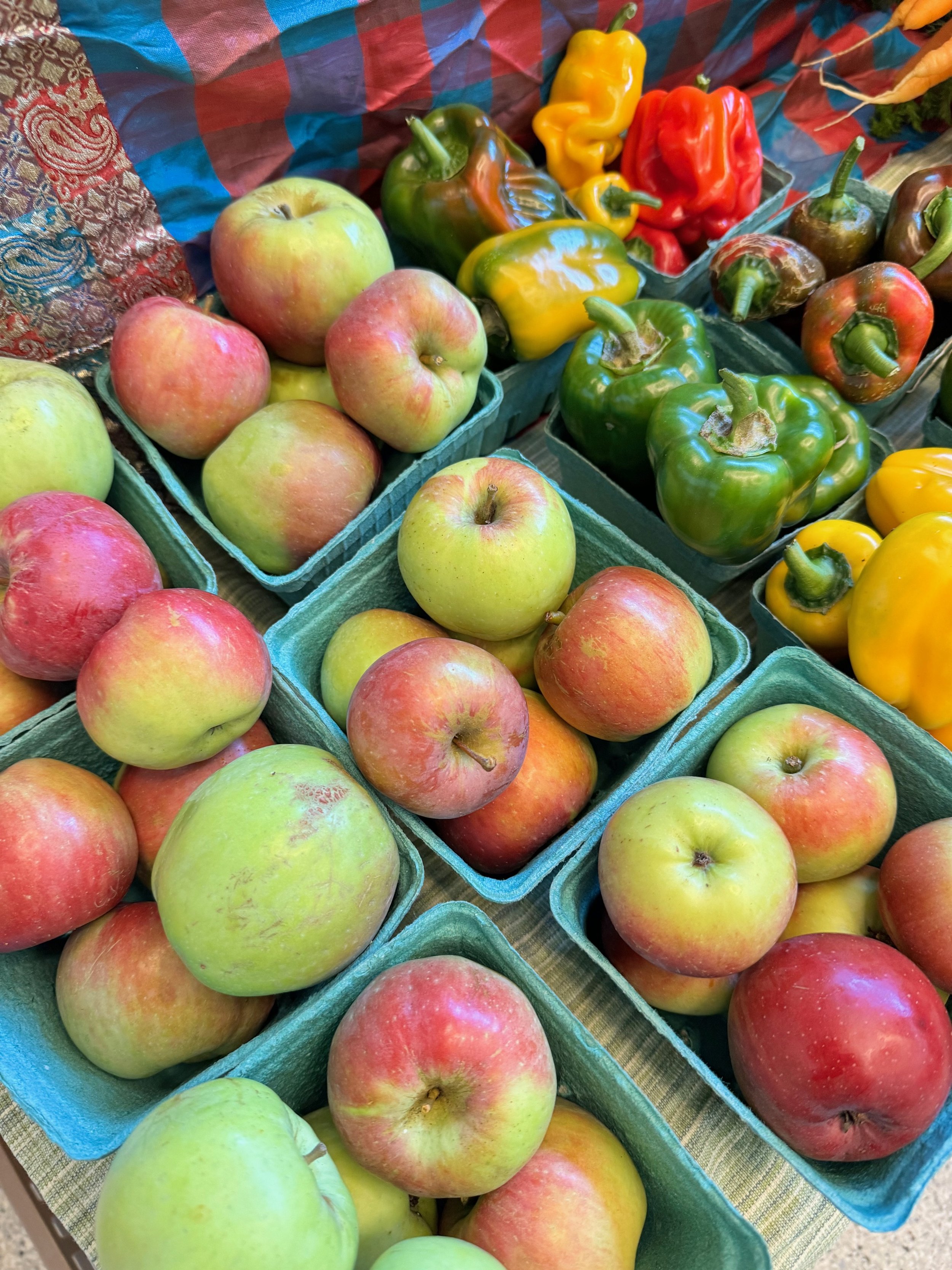 Display of fresh apples and bell peppers in a market stall, with apples in green and red shades and bell peppers in yellow, green, red, and brown colors.