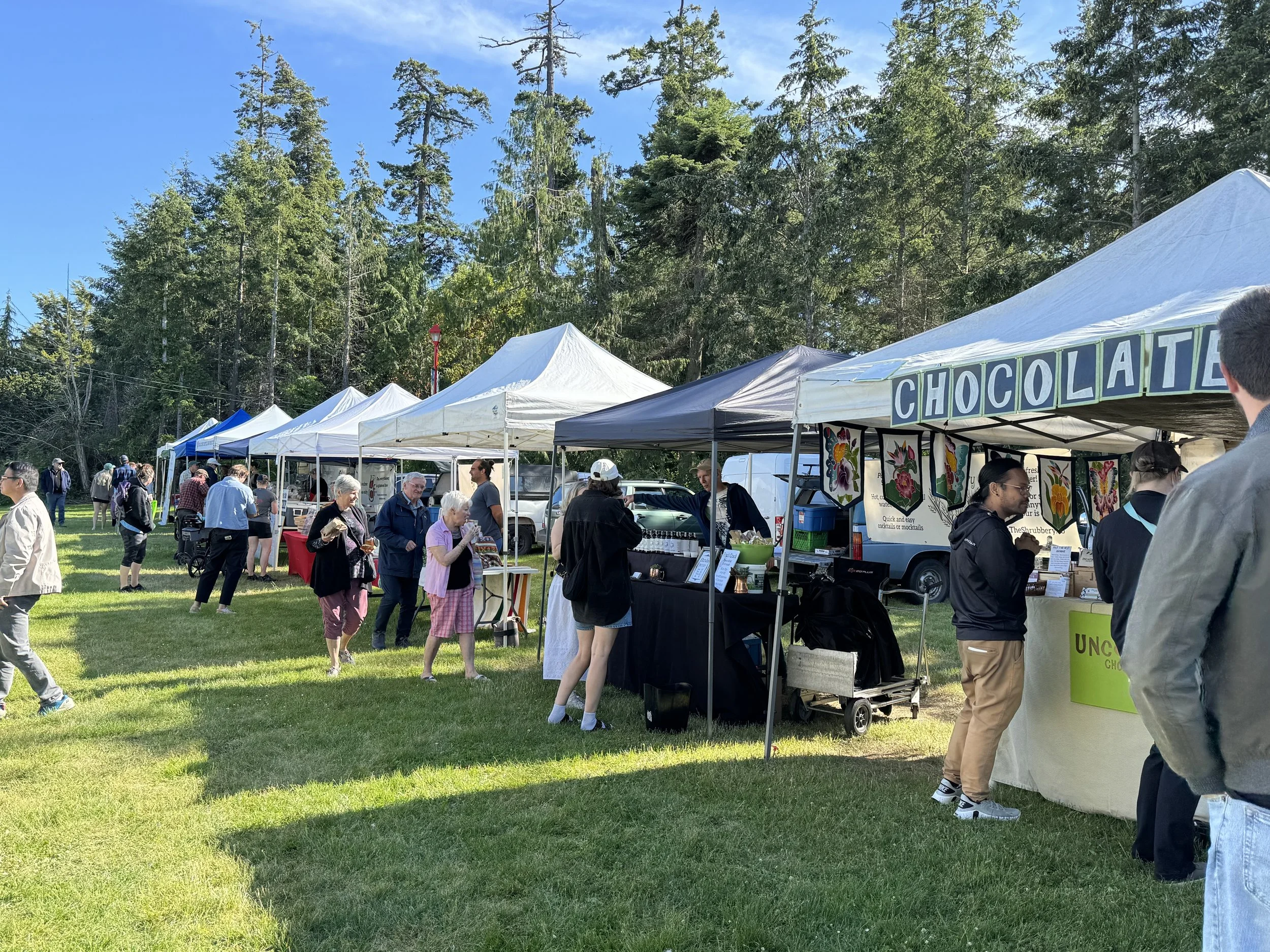 An outdoor market with multiple white tents set up in a grassy area surrounded by tall green trees. Many people are walking around and browsing the stalls.
