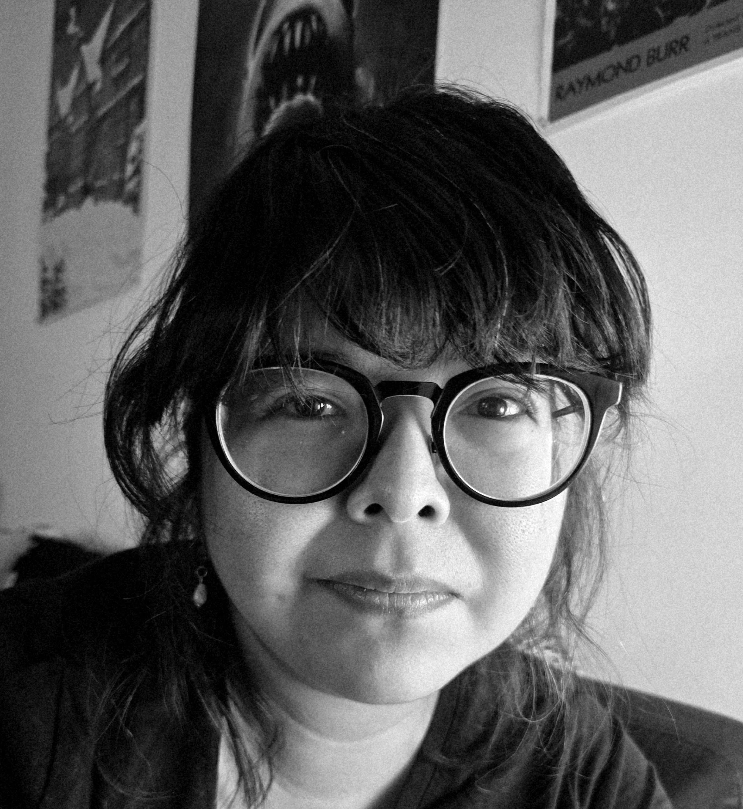 A black and white photo of a woman with dark hair, wearing round glasses, looking into the camera with a slight smile. There are posters on the wall behind her.