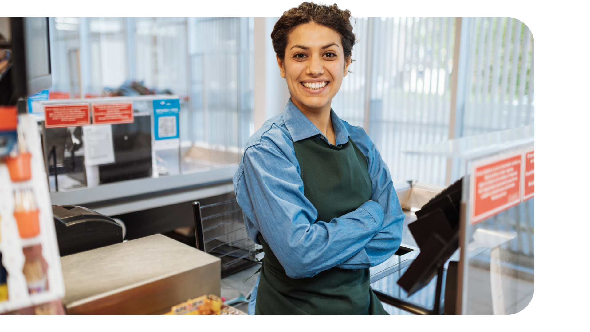 Servicemedarbeider i blå skjorte og forkle på butikk eller kiosk som holder armer i kors og smiler.