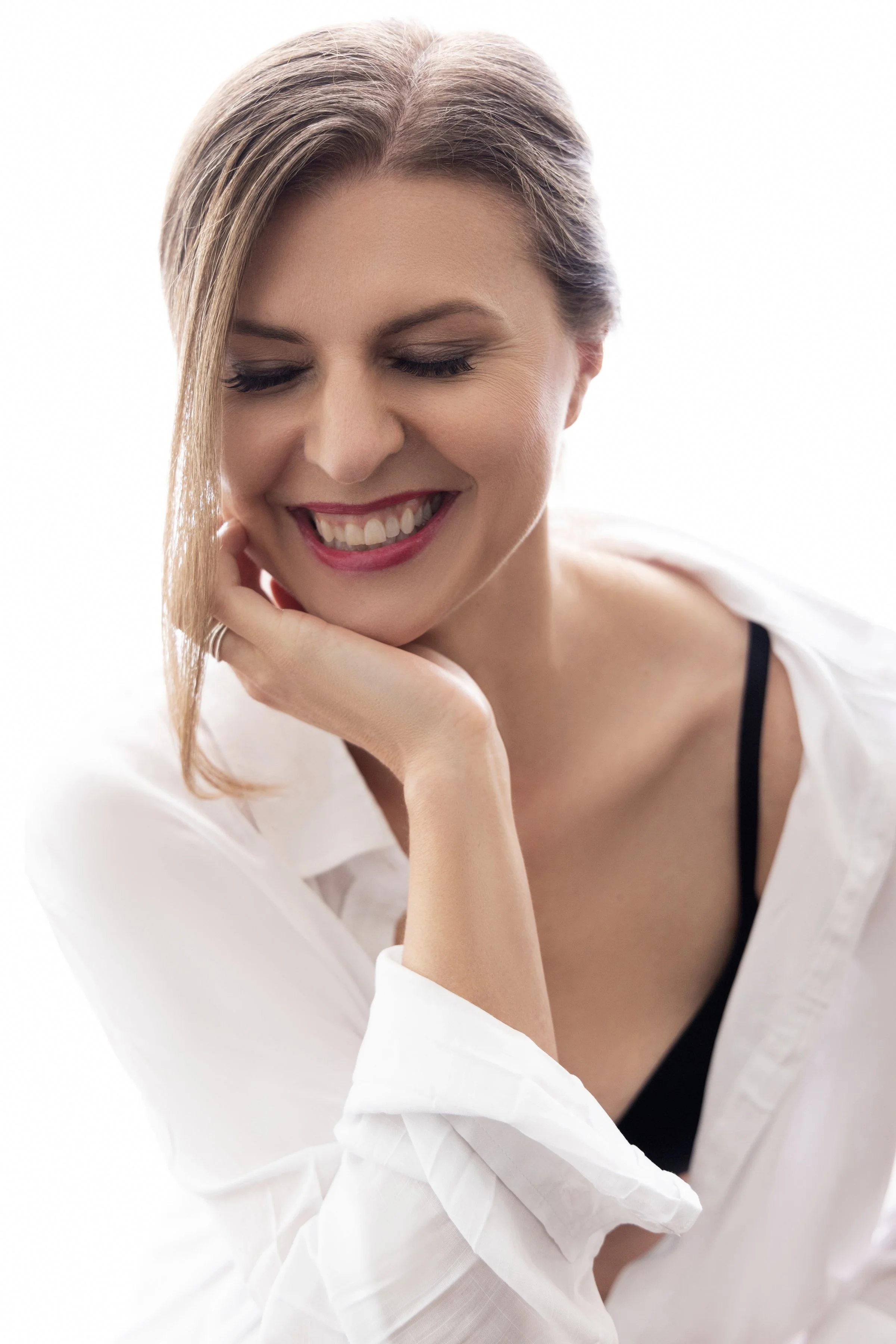 A woman with short hair smiling softly with her eyes closed, wearing a white shirt and black tank top, resting her chin on her hand.