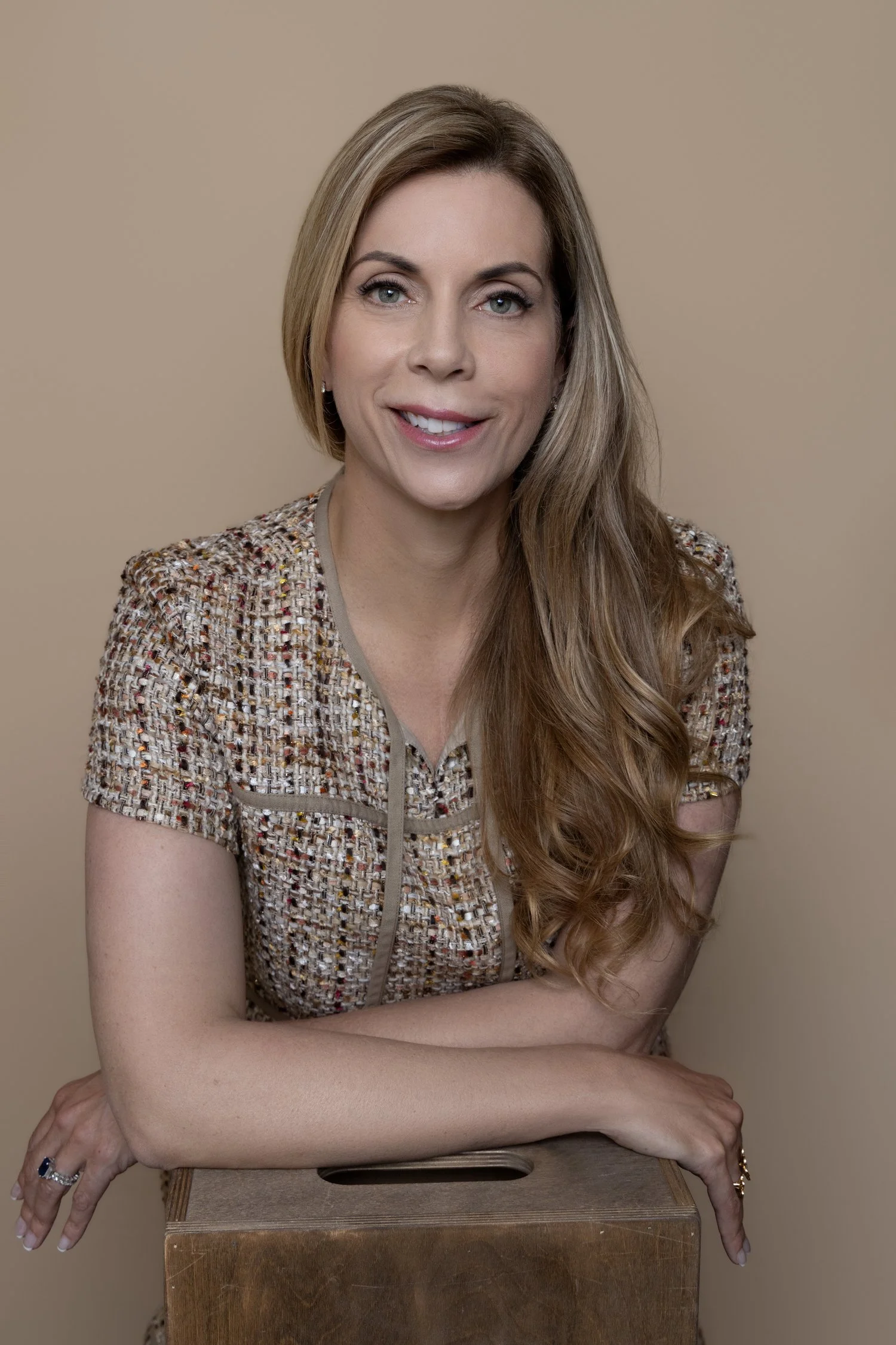 A woman with long, wavy, reddish-brown hair smiling and looking at the camera, wearing a patterned short-sleeved top, posed with her arms resting on a wooden box against a beige background.
