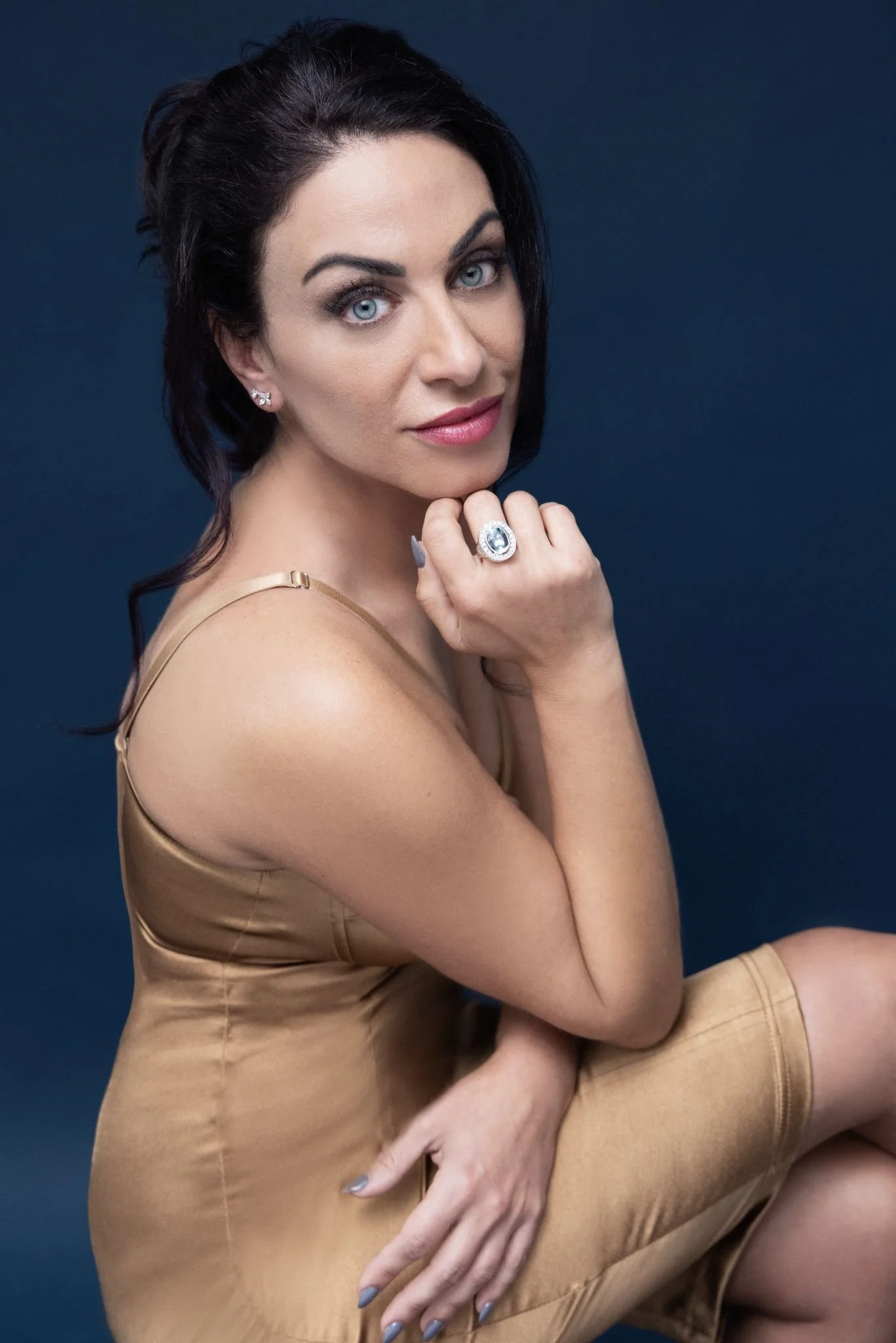 A woman with dark hair, blue eyes, and fair skin poses against a dark background. She is wearing a beige satin dress and large jewelry including earrings and a ring. She is resting her chin on her right hand, with her left hand on her knee, looking at the camera.