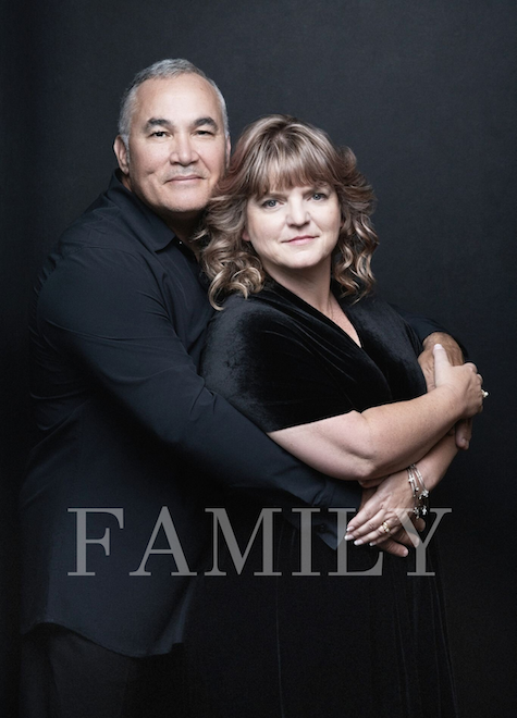 A middle-aged man and woman standing closely against a dark background, with the word 'FAMILY' overlayed at the bottom. The man has short gray hair and wears a black shirt. The woman has shoulder-length wavy hair and wears a black dress, with her arms crossed.