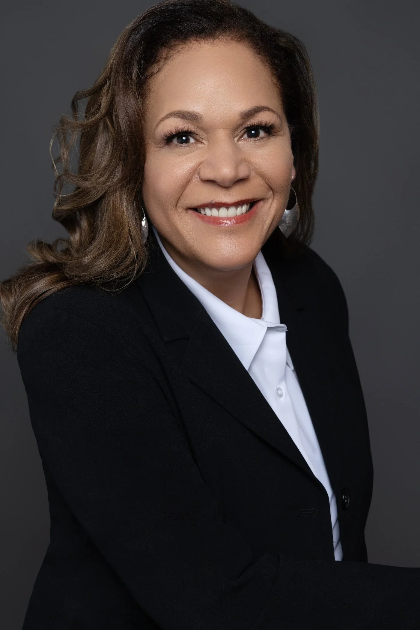 A woman with shoulder-length brown hair, dressed in a black blazer over a white shirt, smiling at the camera against a dark grey background.
