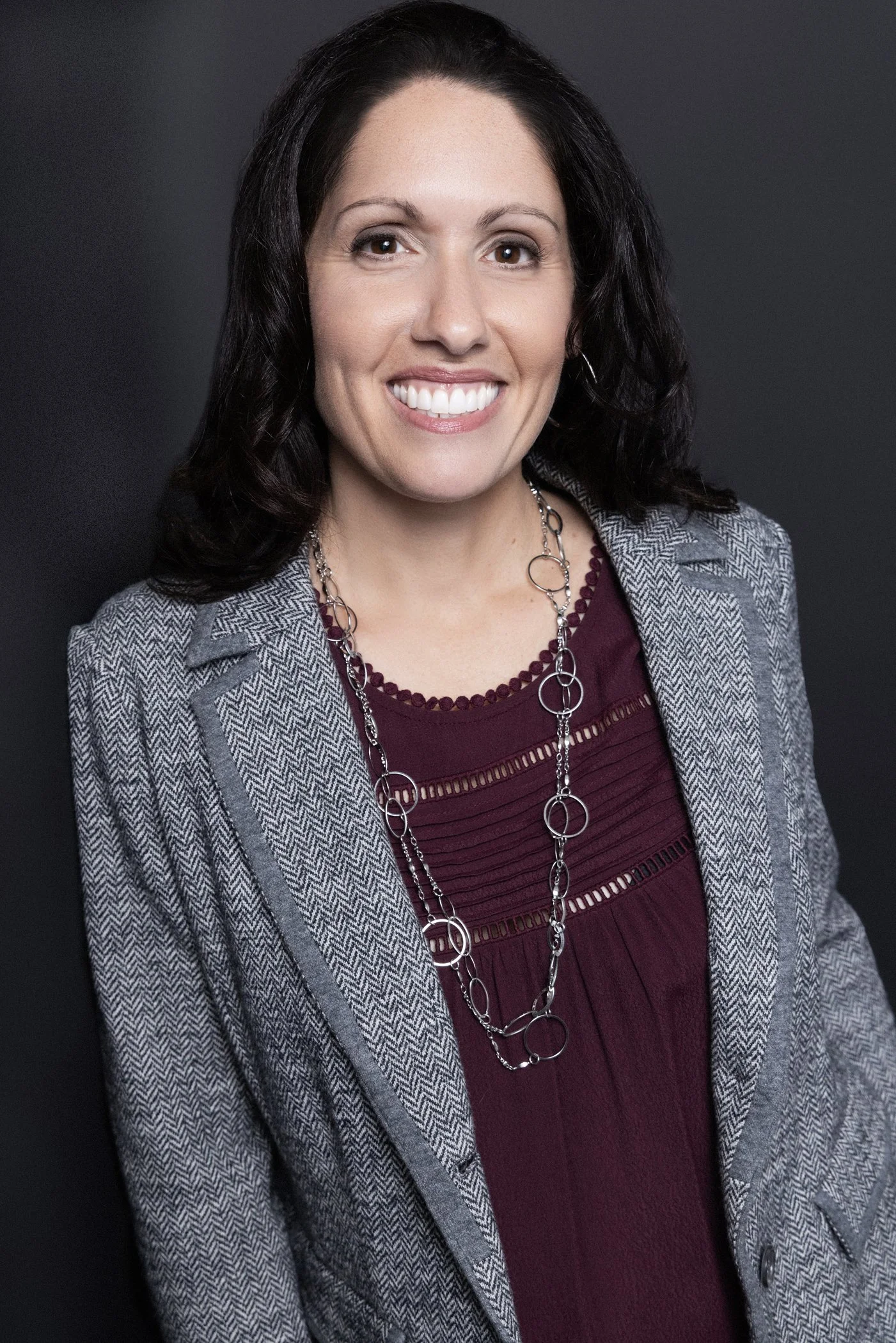 A woman with dark wavy hair, wearing a gray herringbone blazer over a maroon blouse, and accessorized with a long silver chain necklace, smiling against a black background.