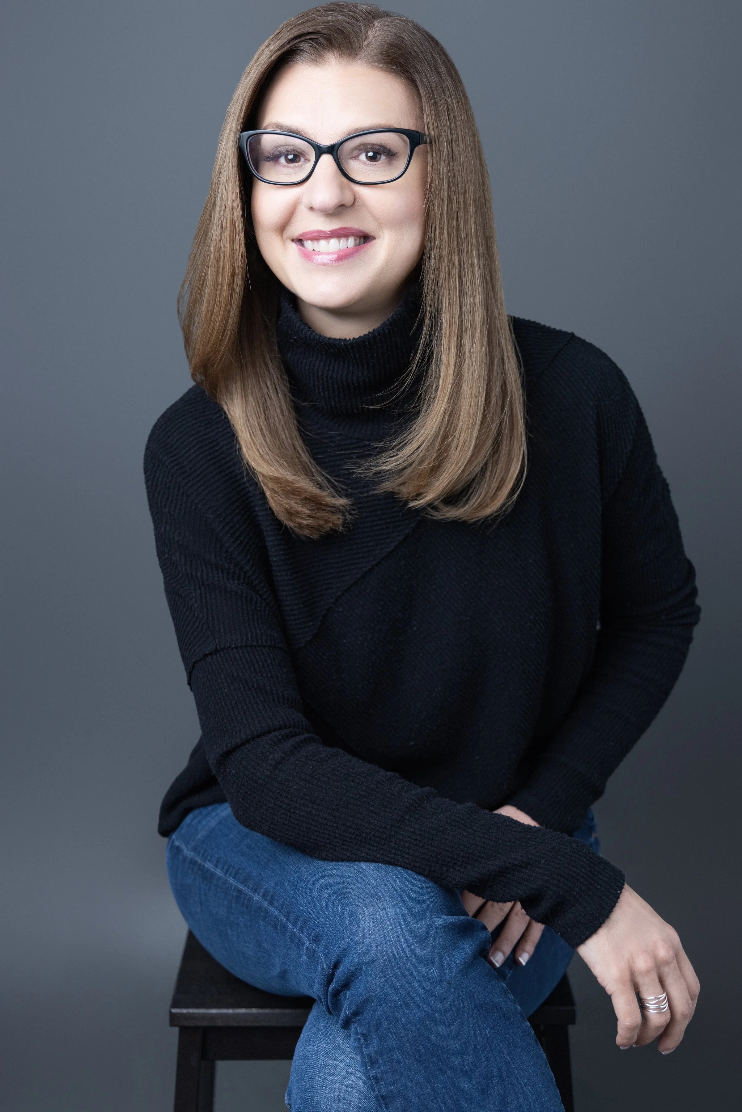 A woman with shoulder-length brown hair, wearing glasses, a black turtleneck sweater, and blue jeans, sitting on a black stool against a gray background, smiling at the camera.