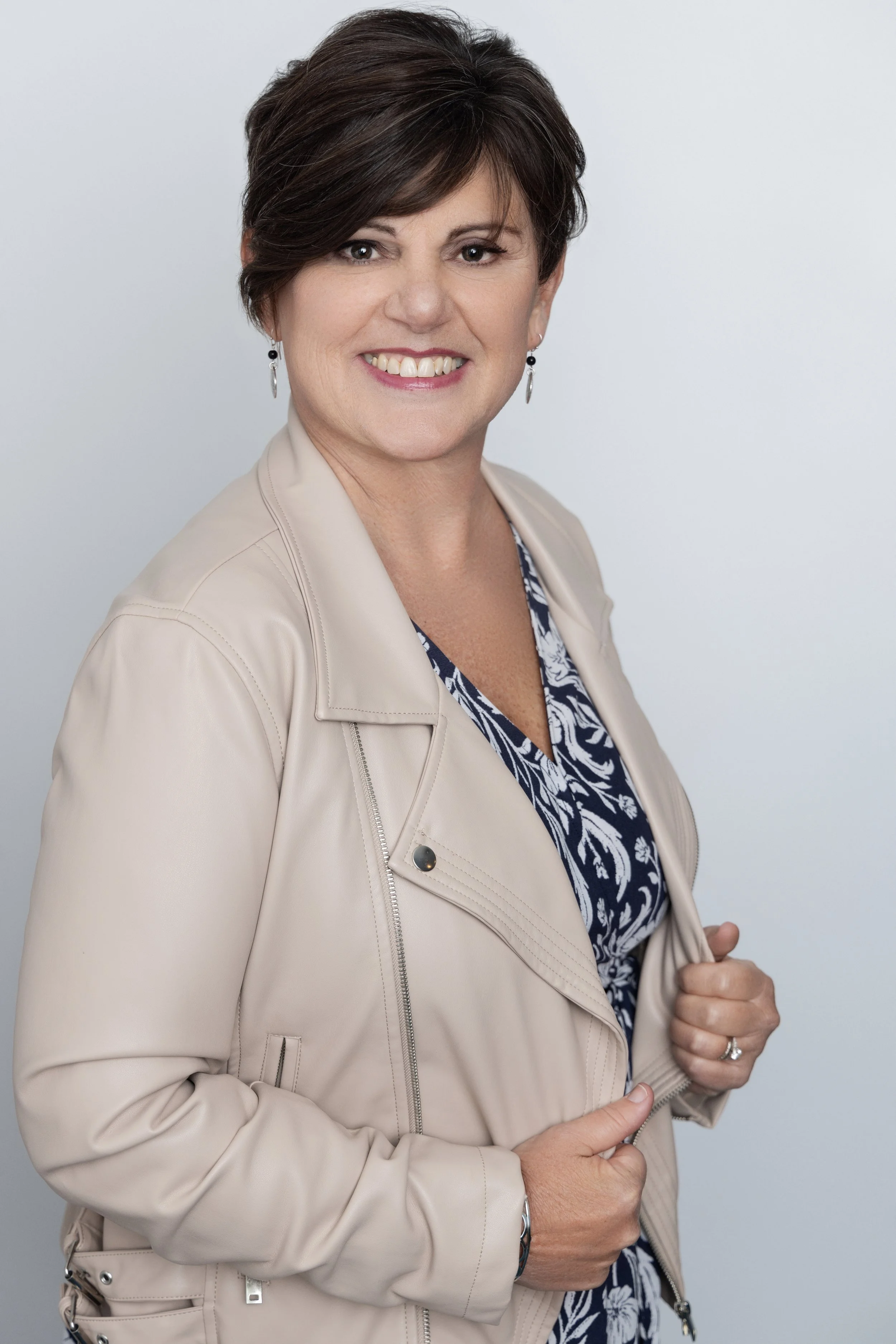 A smiling woman with short dark hair, wearing a beige jacket over a navy and white patterned top, standing against a white background.
