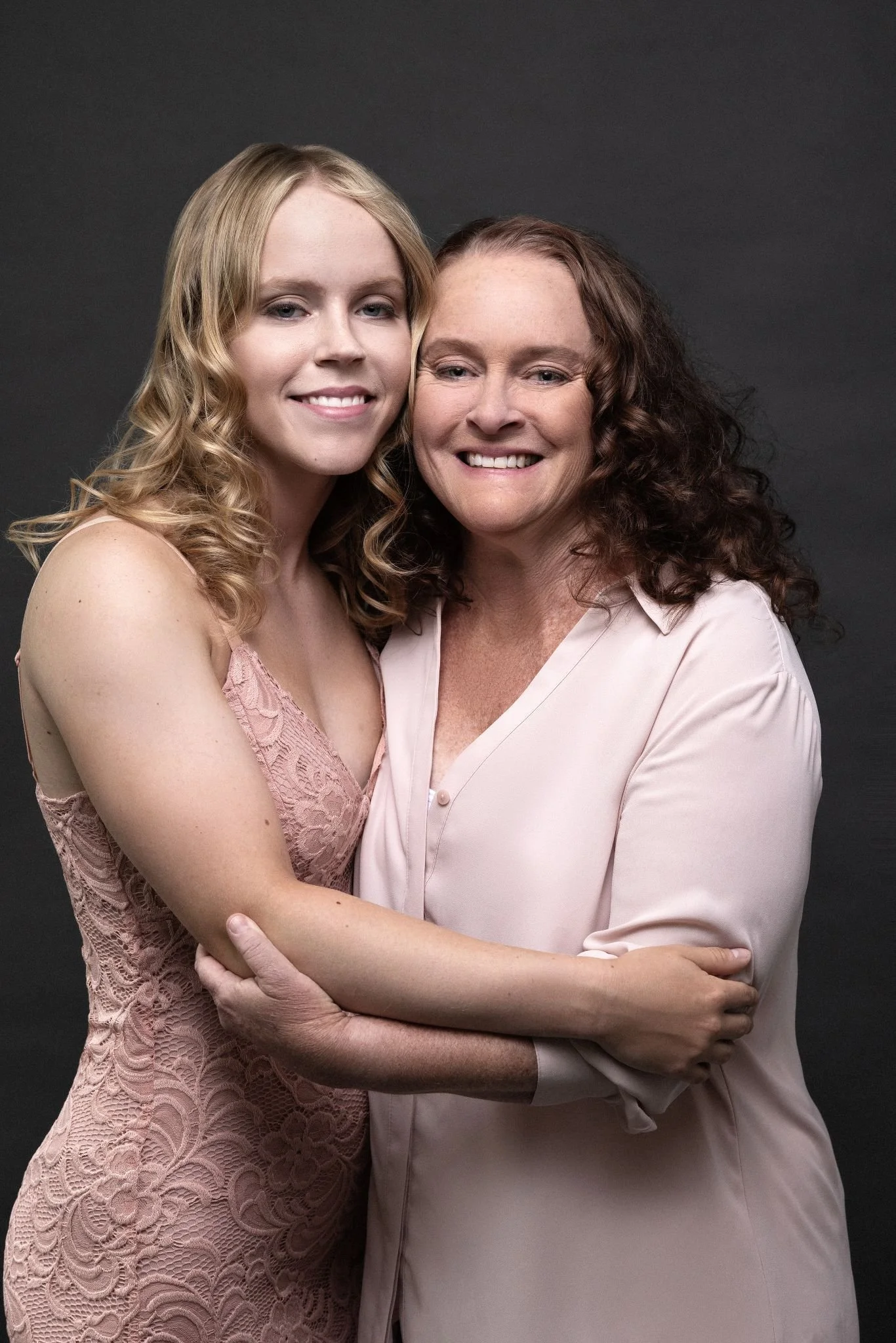 Two women, one younger with blonde hair in a pink lace dress and one older with curly brown hair in a light pink blouse, smiling and hugging in front of a gray background.
