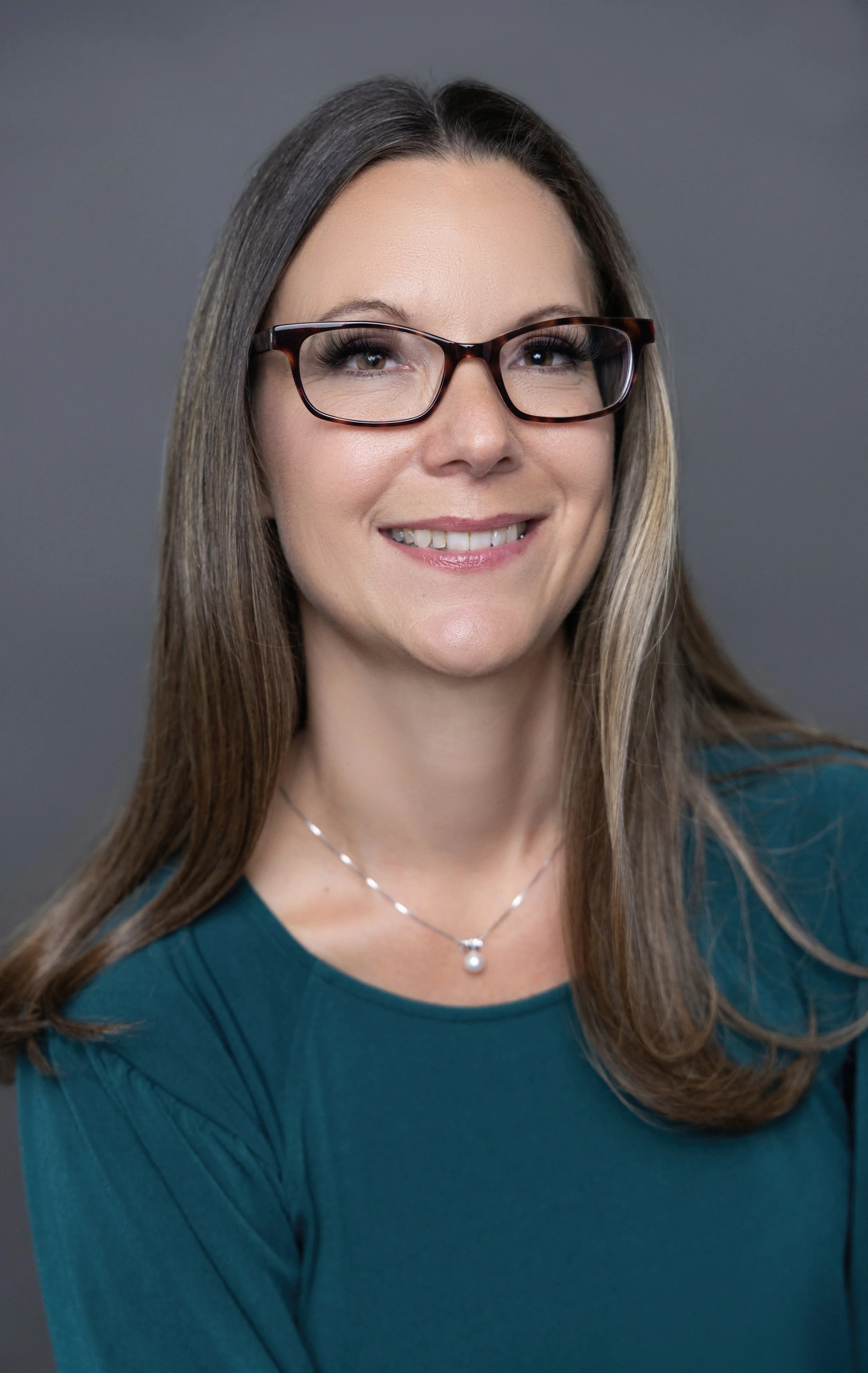 A woman with long brown hair, wearing glasses, a teal top, and a pearl necklace, smiling against a gray background.
