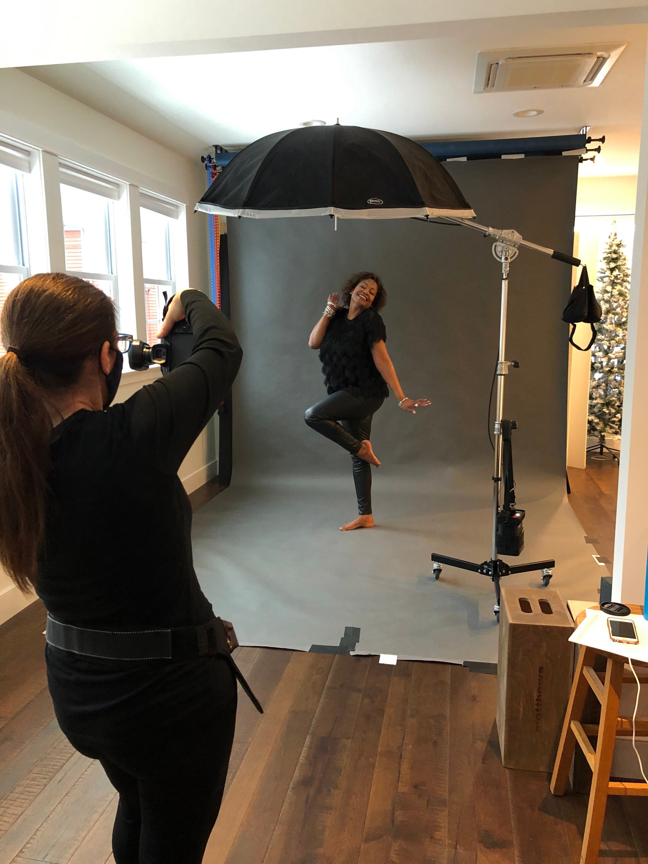 A woman poses playfully while being photographed in a professional photo studio with a gray backdrop and studio lighting equipment.