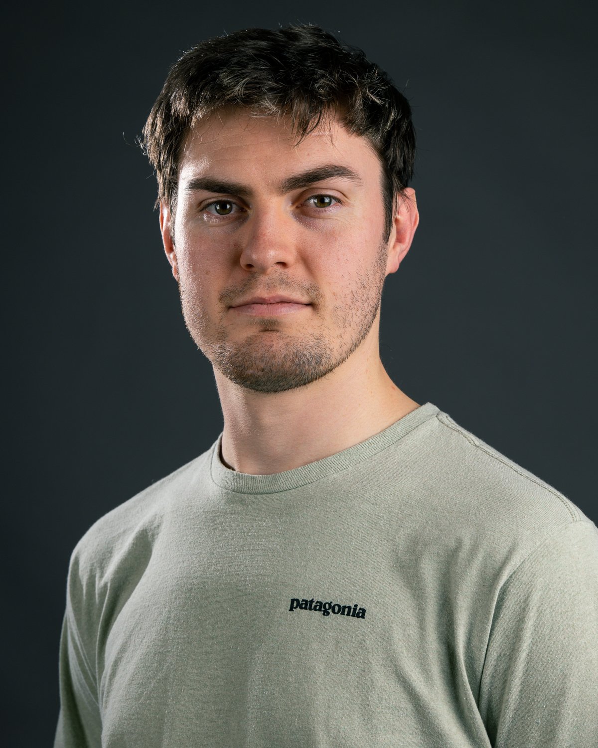 Close-up of a young man with dark hair, green eyes, and light skin, wearing a light-colored Patagonia t-shirt, against a dark background.