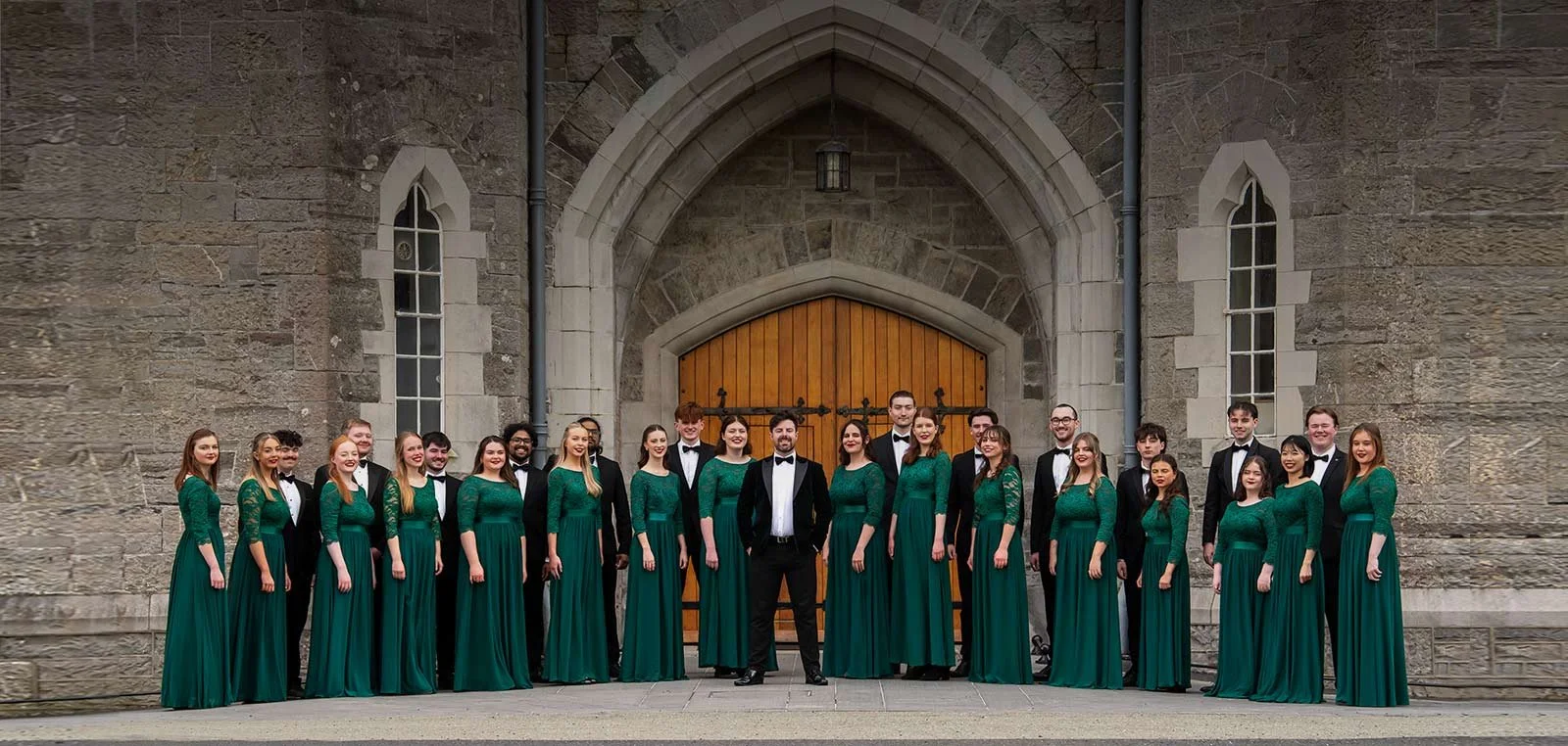 Group of choir singers in green dresses and black tuxedos standing in front of a stone church entrance.