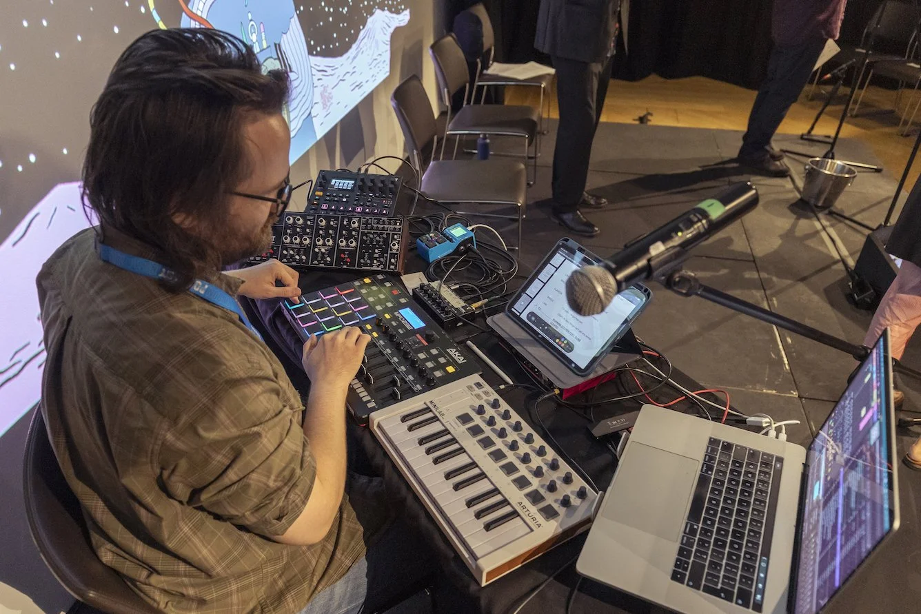 Odinn Orn Hilmarsson with electronic music equipment, including a MIDI controller, synthesizer, and a laptop, at a table on a stage. There is a microphone on a stand in front of him and a tablet displaying a music interface.