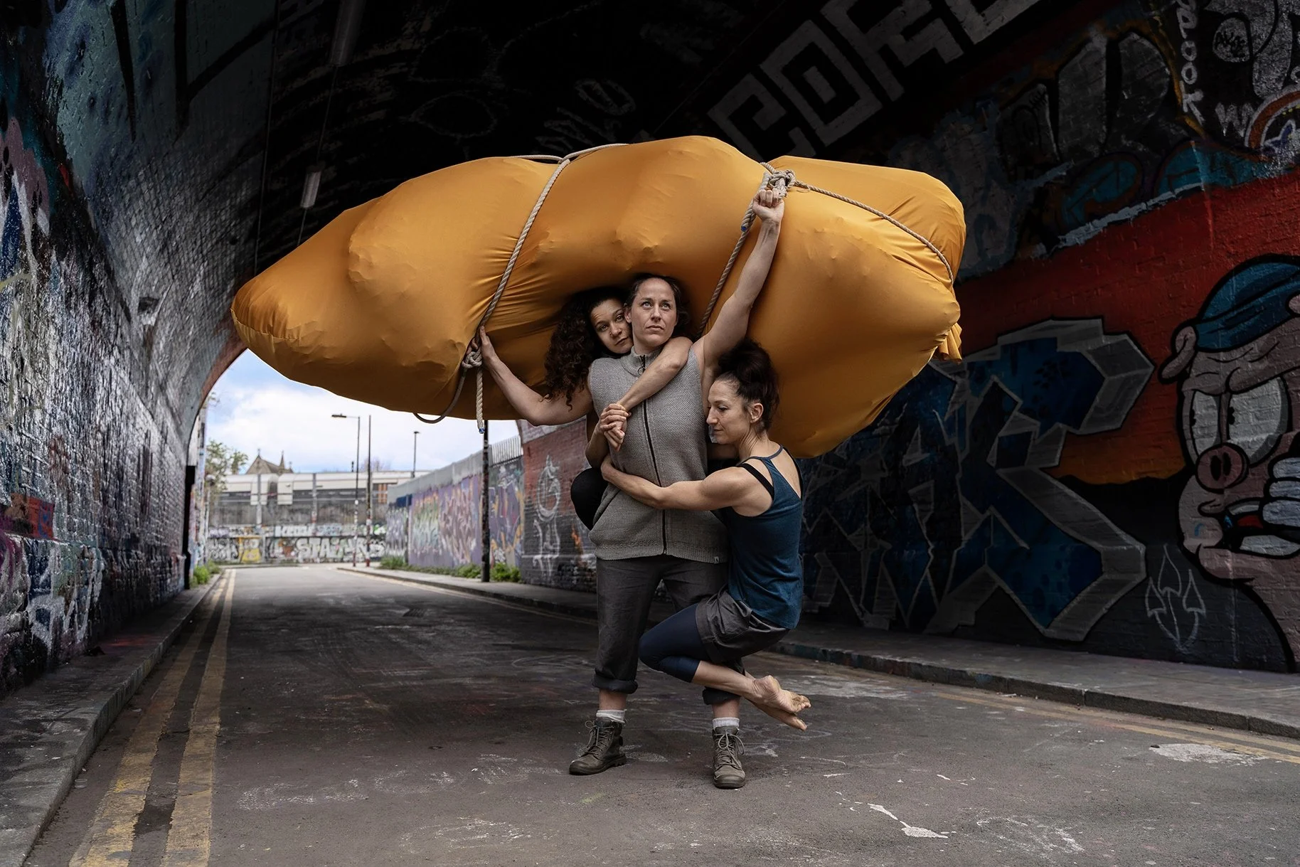 Three women holding up an orange sleeping bag or bundle in a graffiti-covered tunnel.